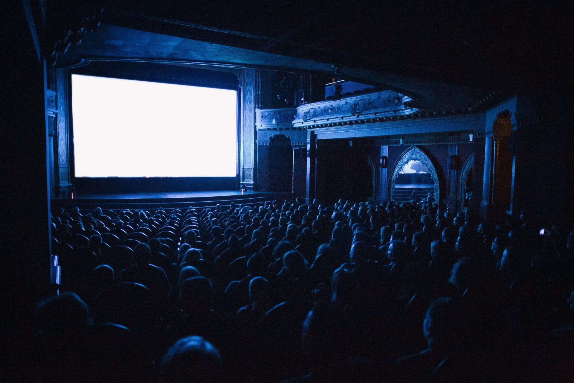 Karanlık bir sinema salonunda izleyiciler parlak beyaz bir ekrana bakıyor. Mavi ışıklandırma, süslü mimariyi vurguluyor.