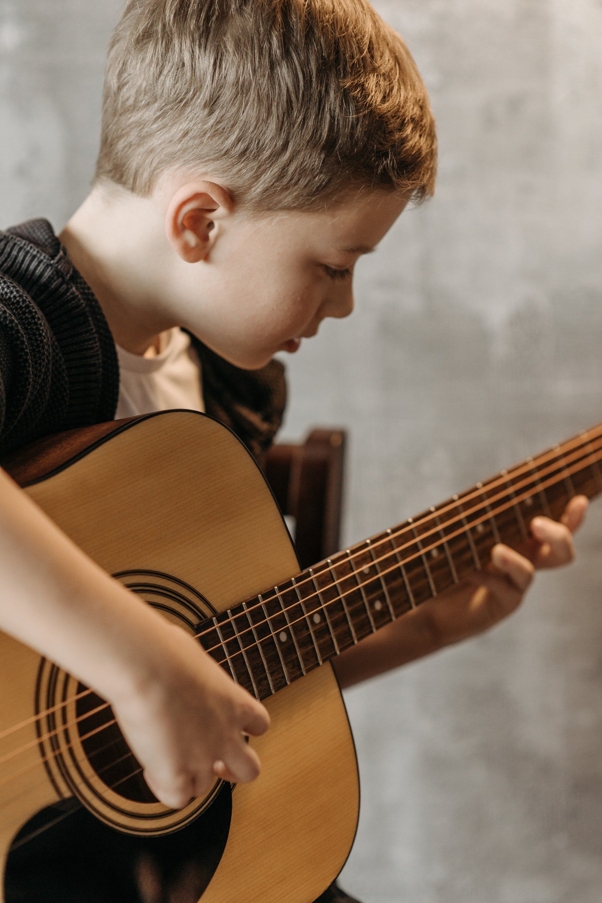 A child playing an acoustic guitar while looking down at the fretboard against a grey wall.