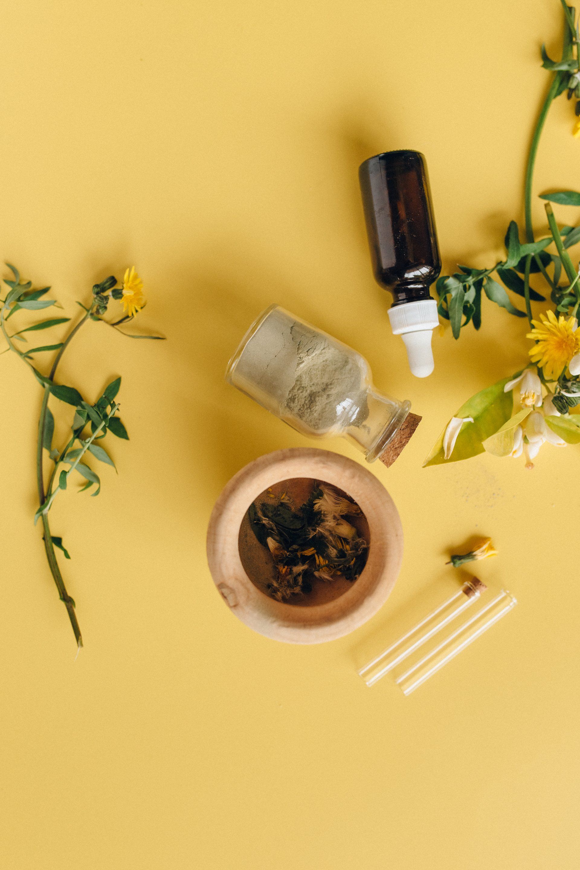 Bottled items with greenery on a yellow background, including bottles, a bowl, and cotton swabs