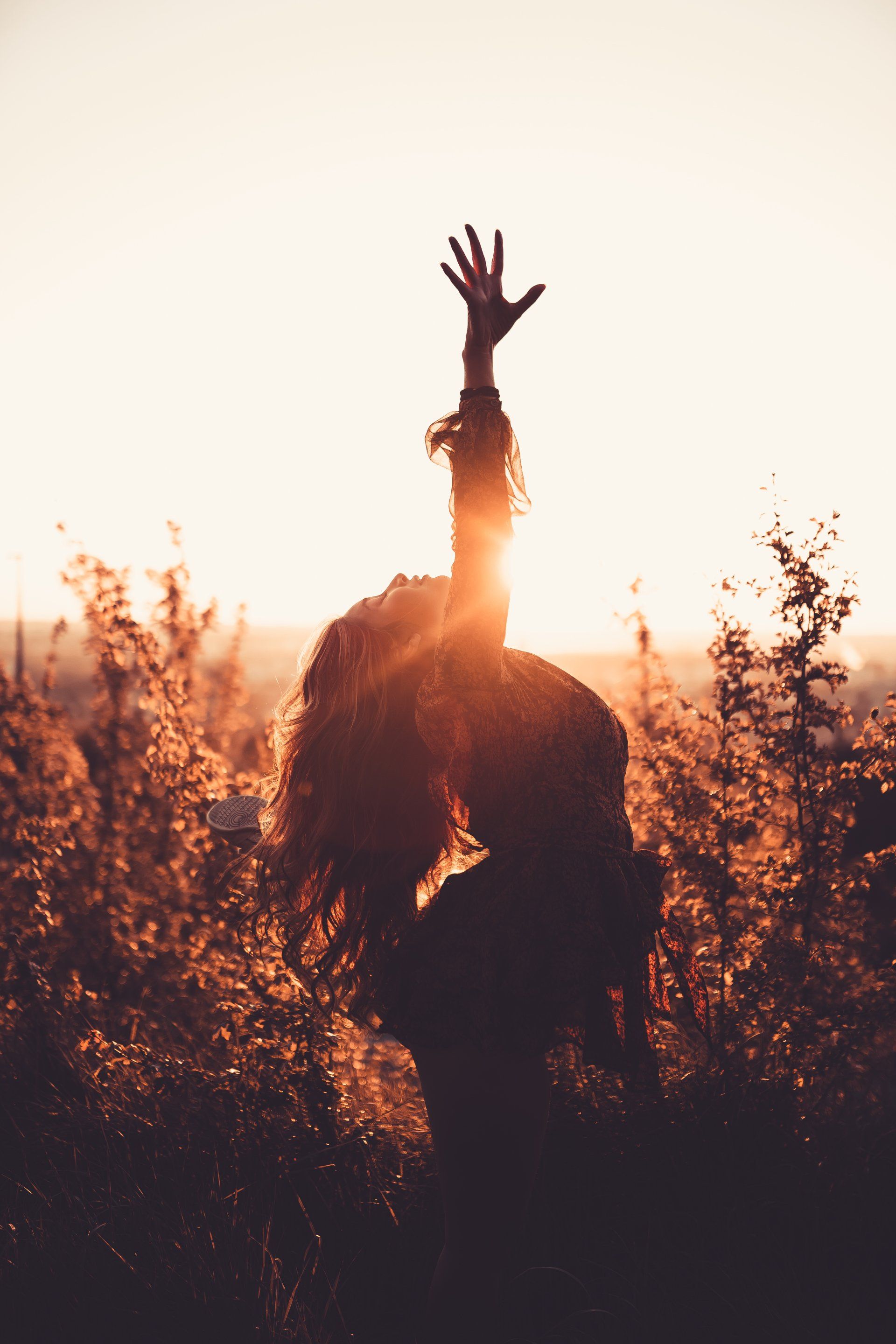 Person raising arm toward setting sun amidst tall grass; silhouetted in golden light.