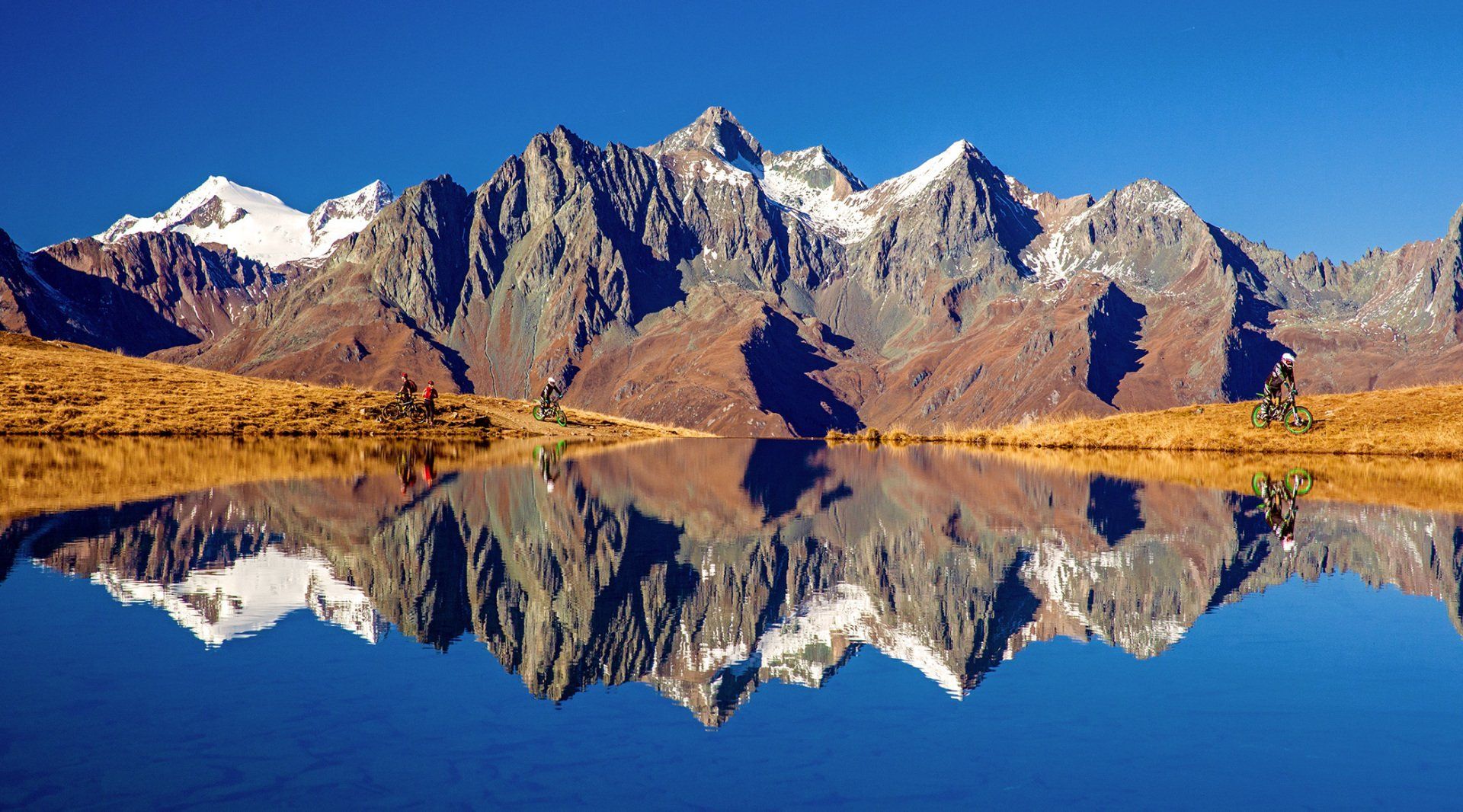 Schneebedeckte Bergketten spiegeln sich in einem stillen blauen See, umgeben von goldbrauner Vegetation unter klarem blauen Himmel.