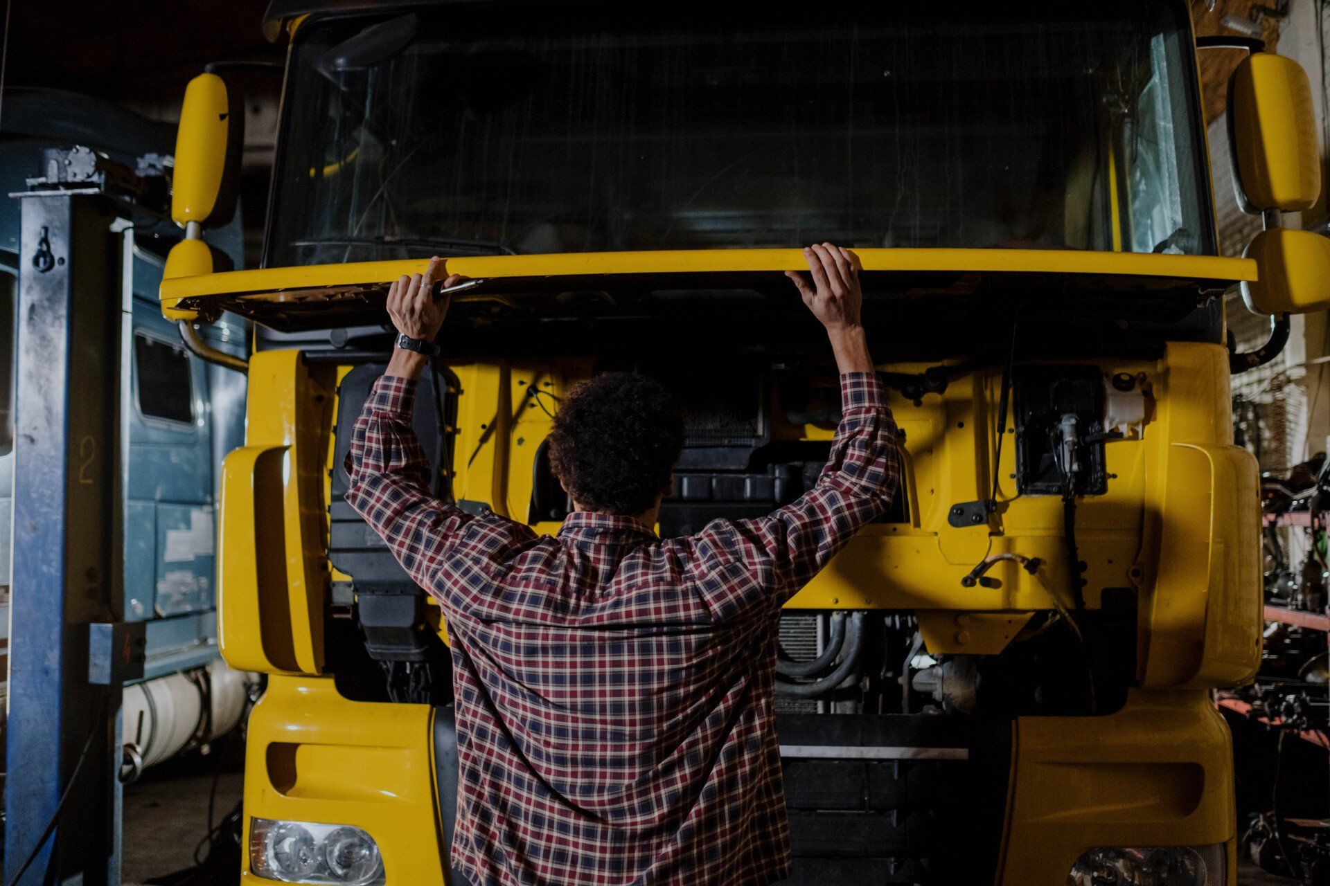 Mechanic in plaid shirt working on yellow truck engine in garage.