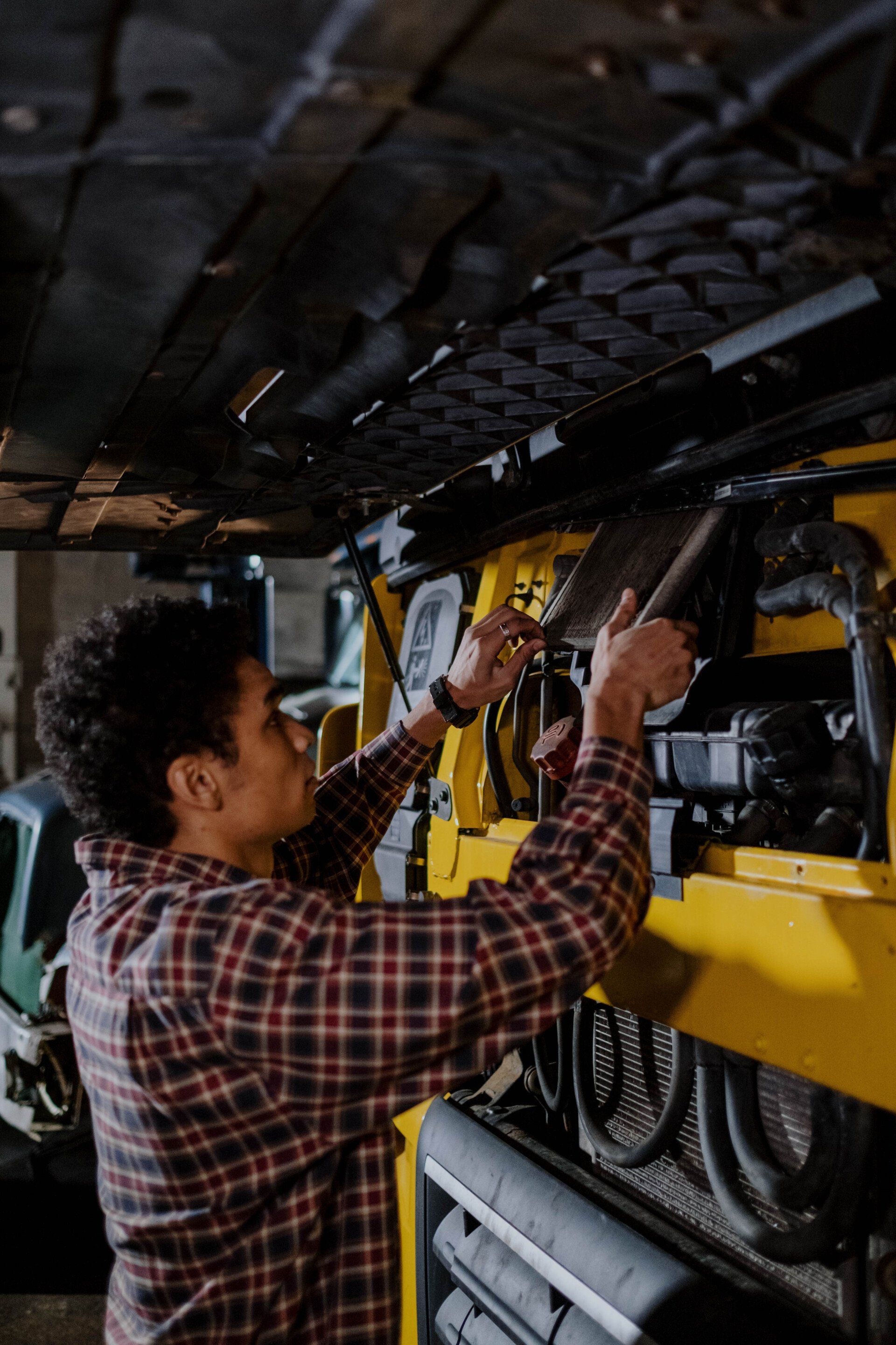 A person wearing a plaid shirt works on the engine of a yellow construction vehicle in a dimly lit garage.