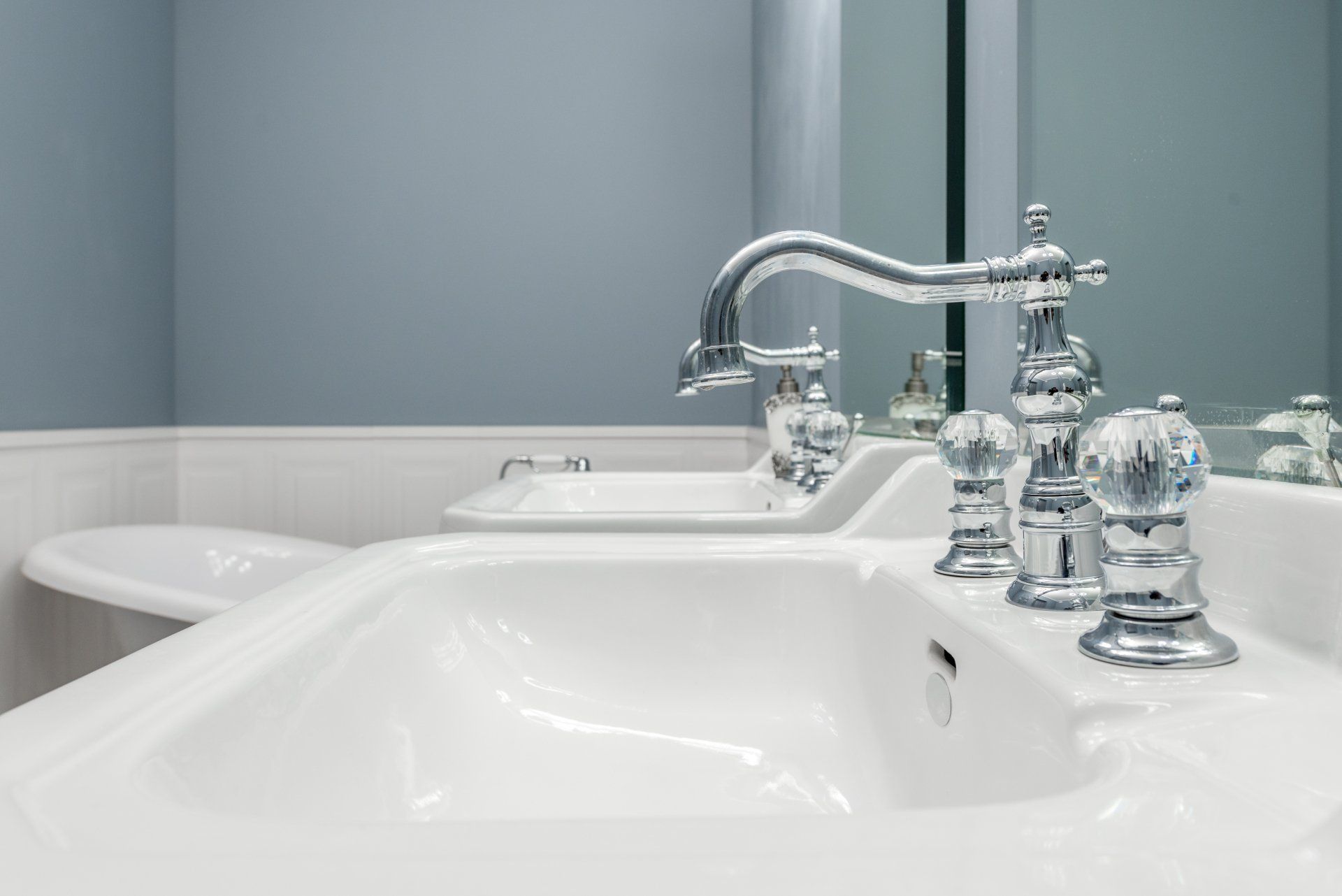 Close-up of a white bathroom sink with ornate chrome faucet and crystal knobs. A second sink is in the background.