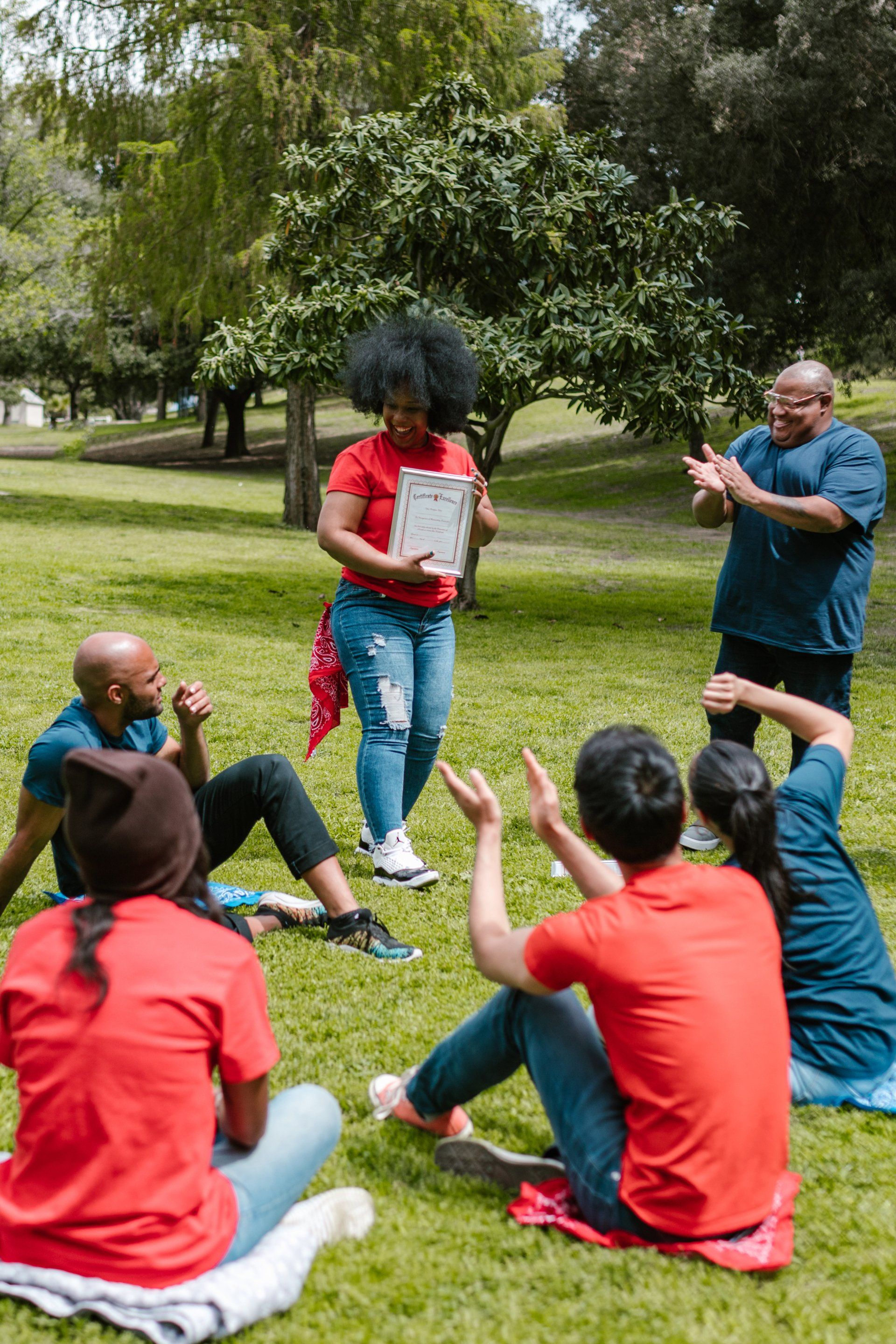 Group of people in a park; one woman presents a paper, others clap and sit on the grass.