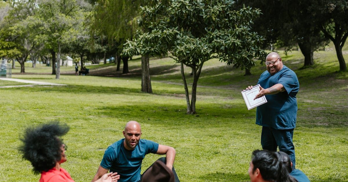 People sitting on grass in a park while a man in a blue shirt stands holding papers.