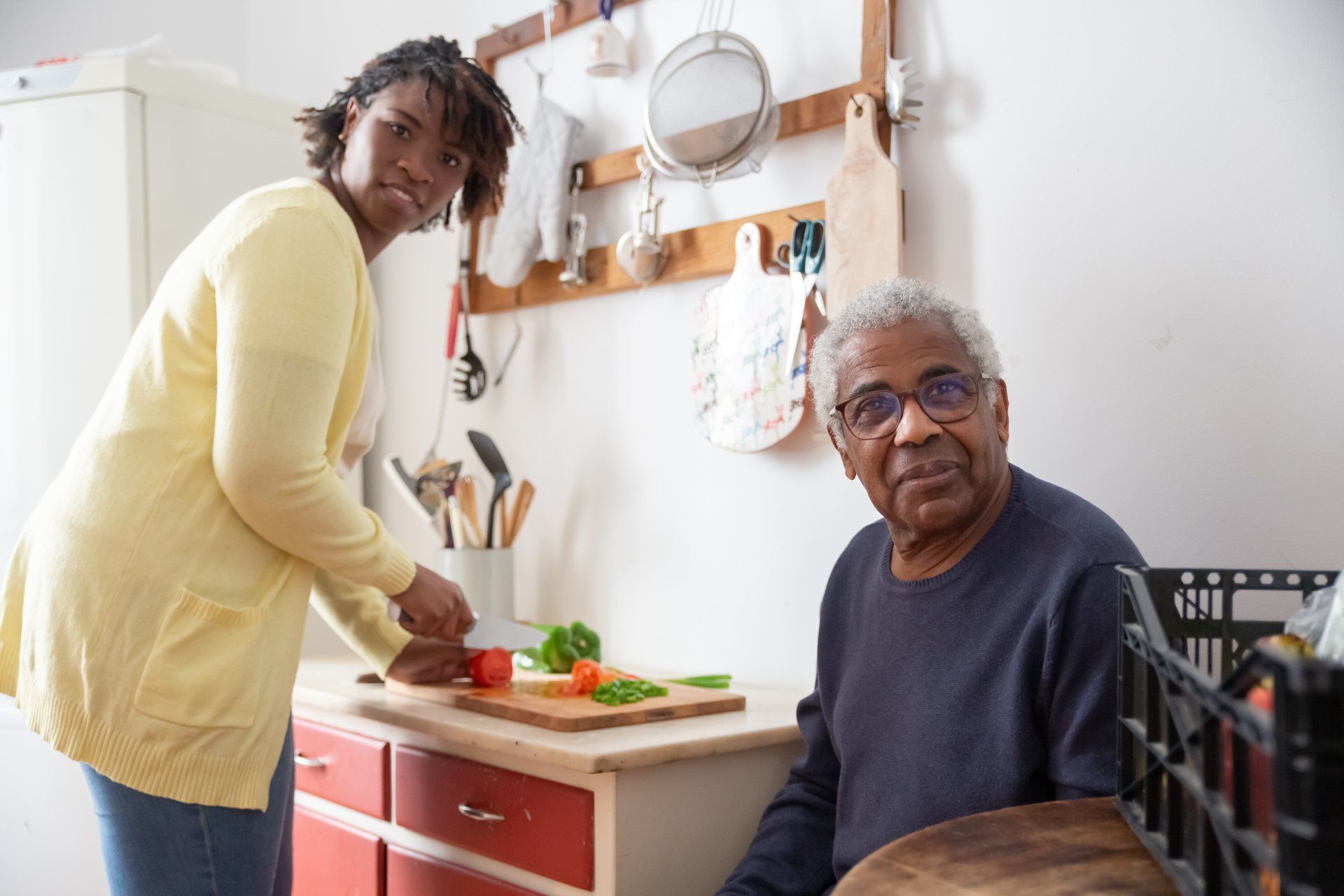 Two people prepare food in a bright kitchen, one chopping vegetables while the other sits nearby.