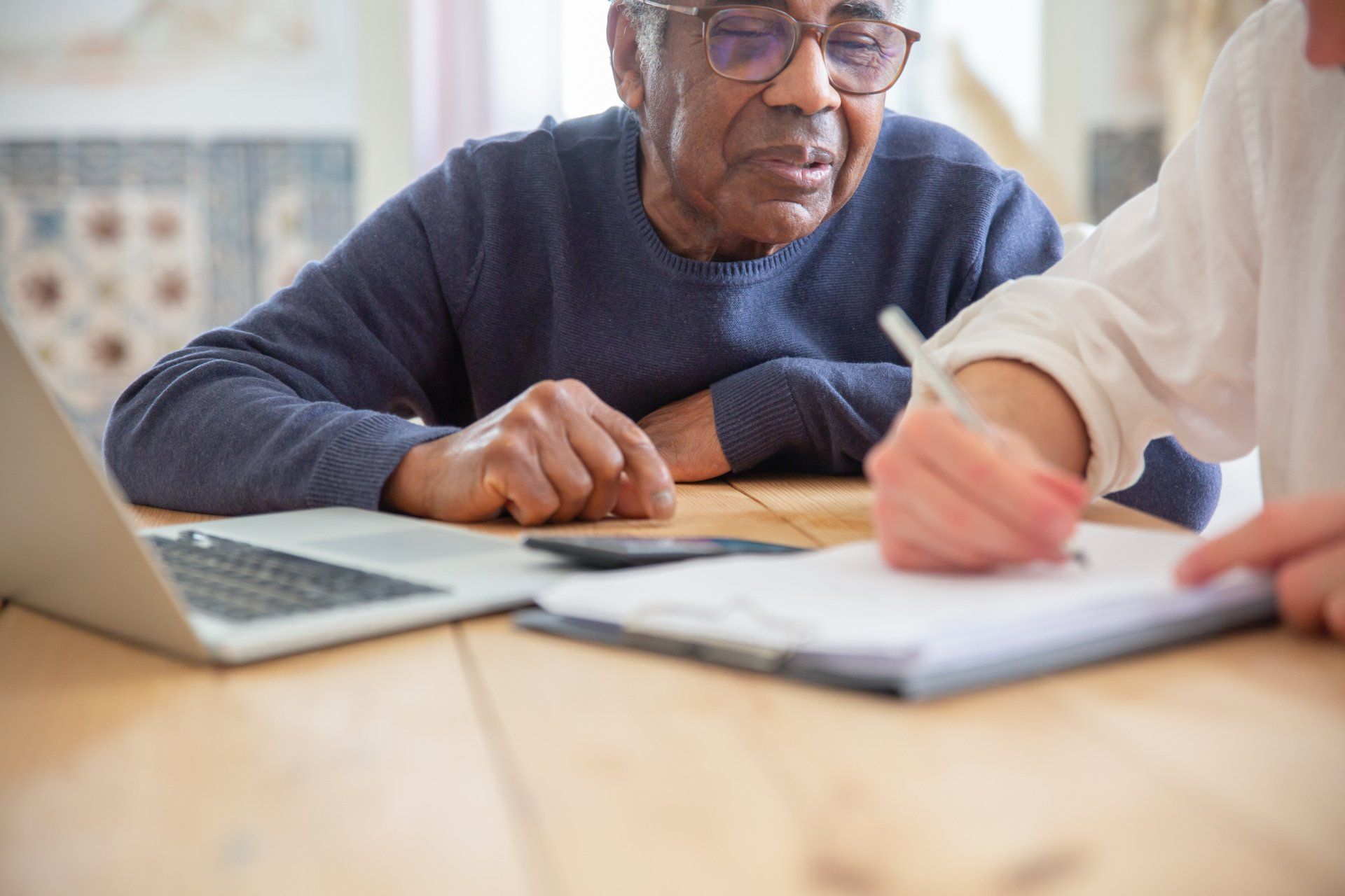 An elderly man is sitting at a table with a laptop and a clipboard.