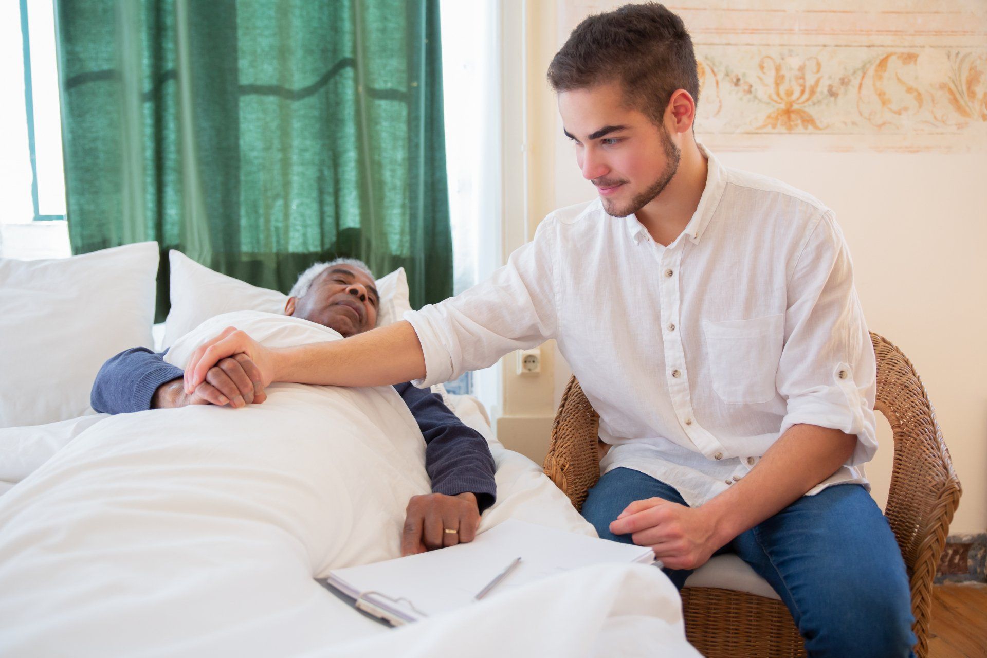 Young man comforting elderly person in bed, holding hands. They are in a room with a window and wicker chair.