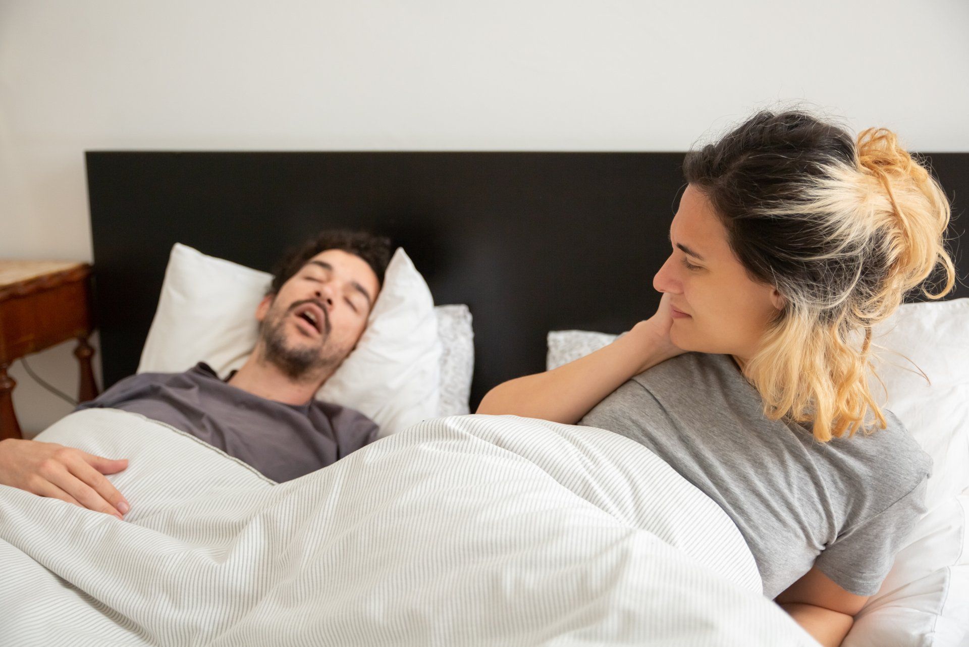 Woman in bed watches man sleeping; he's snoring. Bedroom setting with white linens and black headboard.