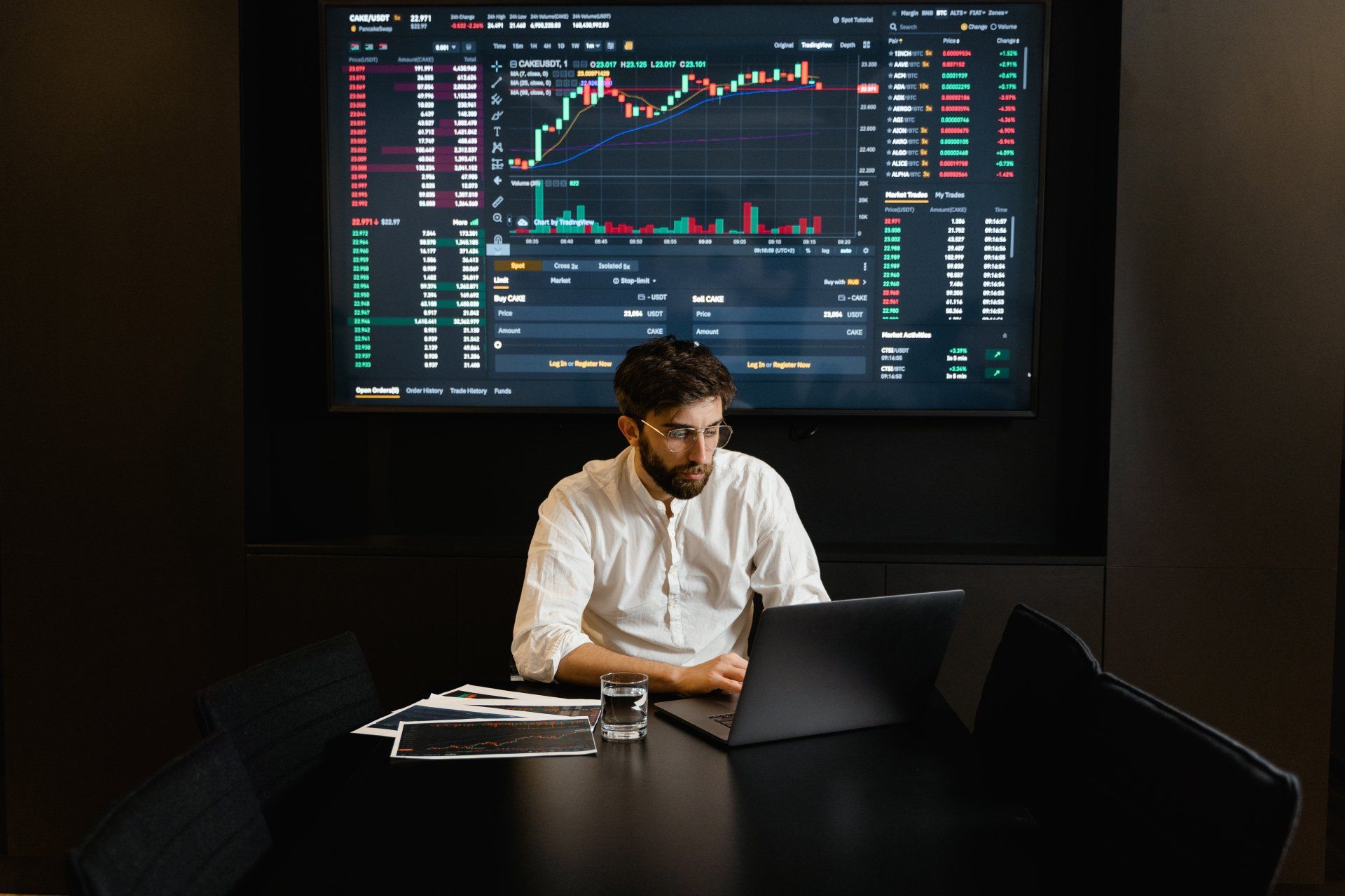 Man working on a laptop in front of a large stock market trading screen in a dark room