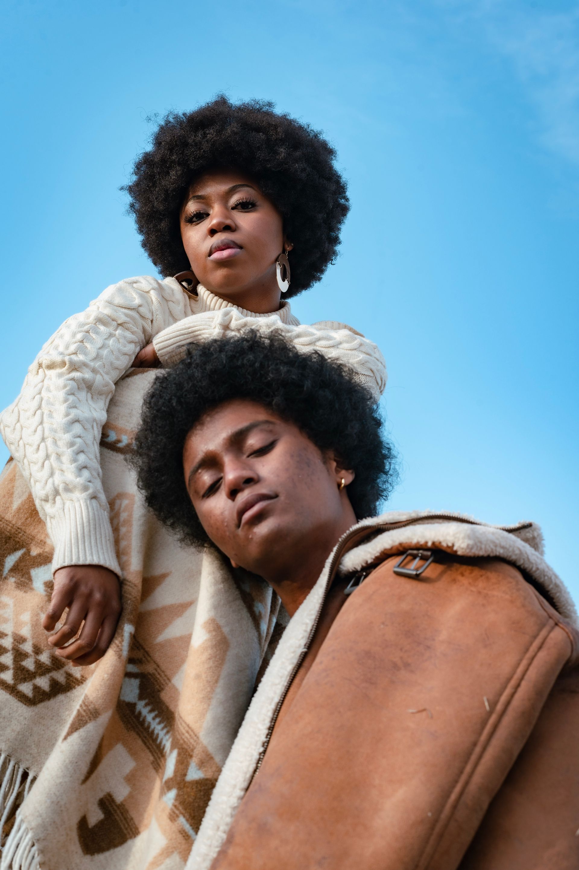 Two people against a blue sky. One draped in a blanket, the other wearing a sweater and looking towards the viewer.