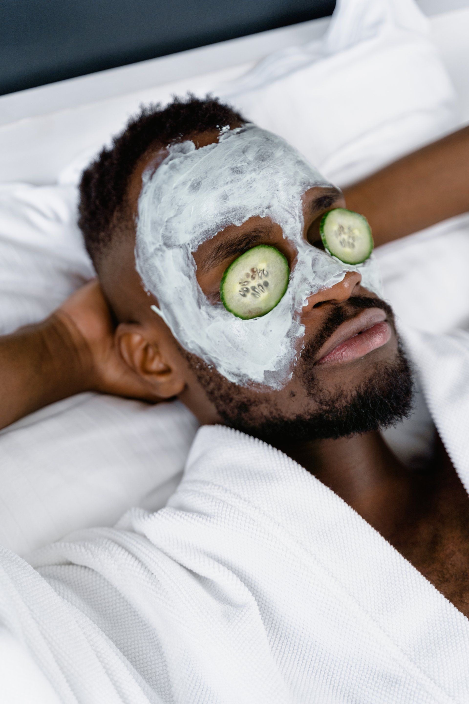 Een man ligt in bed met een masker op zijn gezicht en komkommers op zijn ogen.