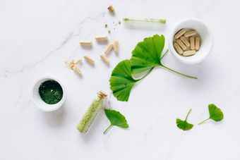 Green leaves and herbal capsules arranged on a white background with small bowls of powder and pills