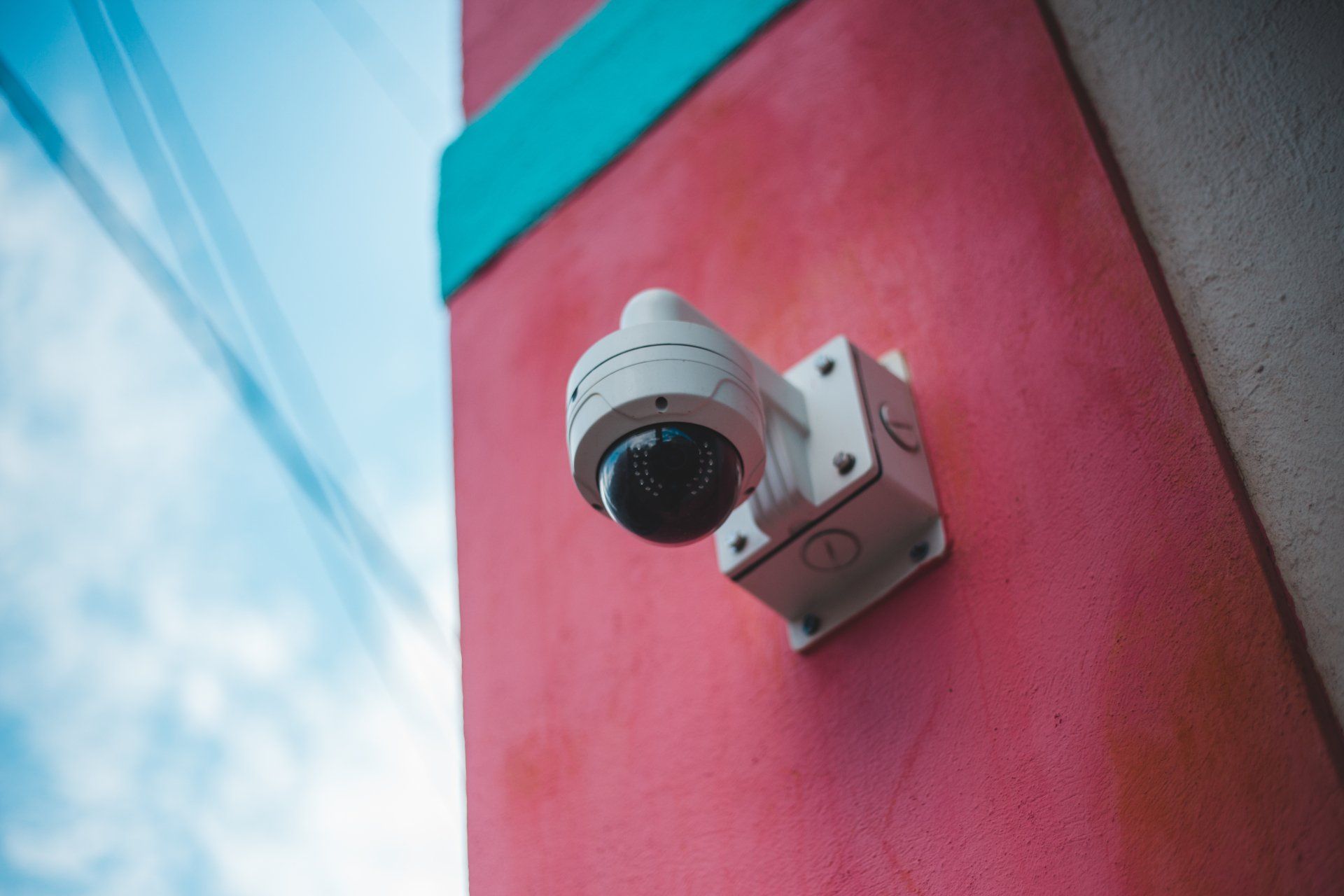 White security camera mounted on a pink building against a blue sky.