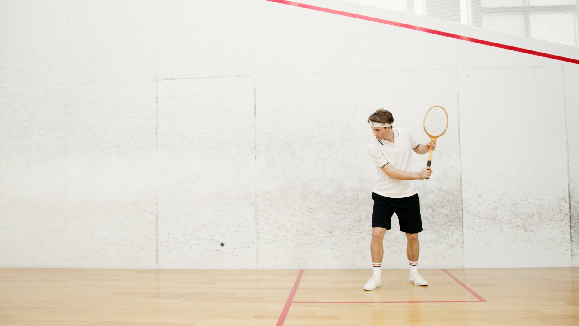 A person in a white shirt and black shorts prepares to hit a squash ball with a racket on an indoor court.