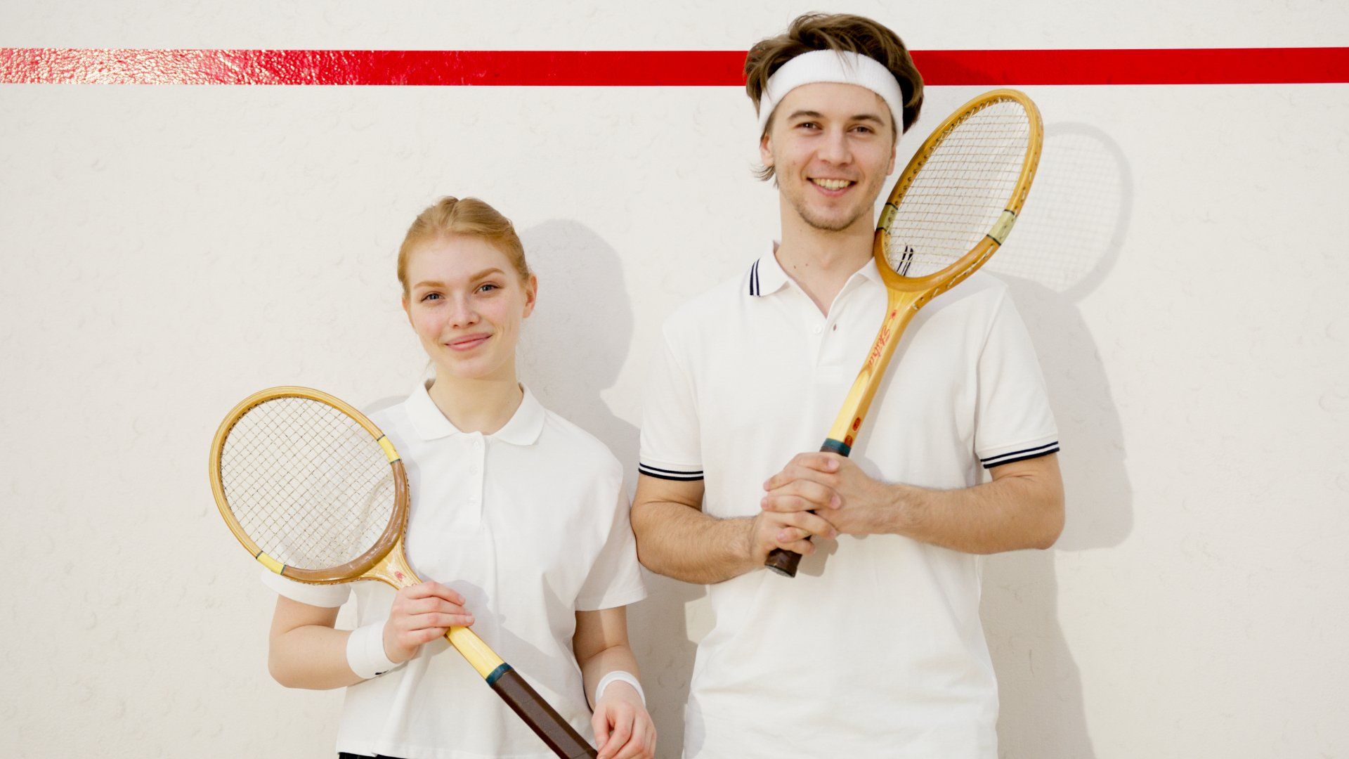 A smiling man and woman wearing white squash outfits and holding wooden rackets stand in front of a white court wall.