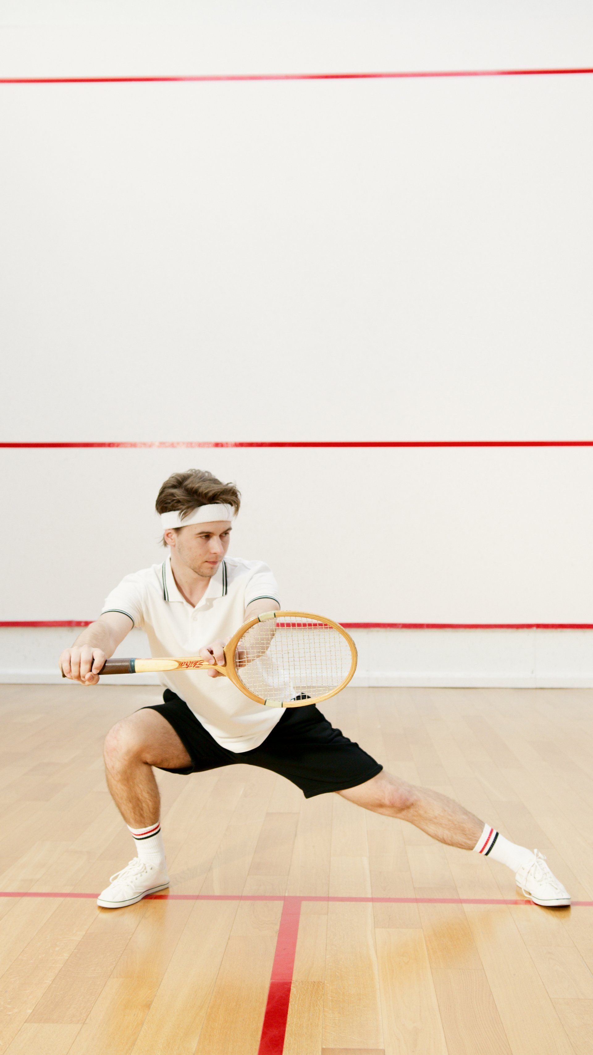 A player in white sportswear and a headband performs a deep lunge while holding a squash racket on a wooden court.