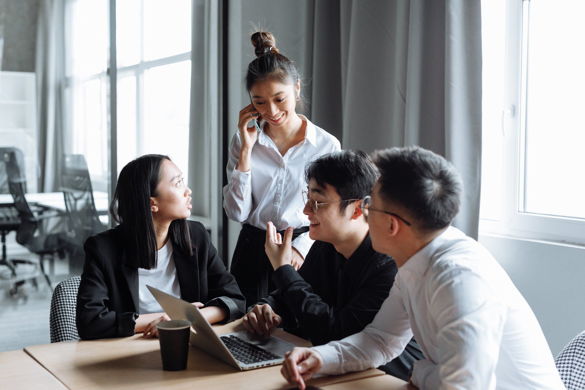Four people collaborating around a laptop at a table in an office setting. One person on the phone.
