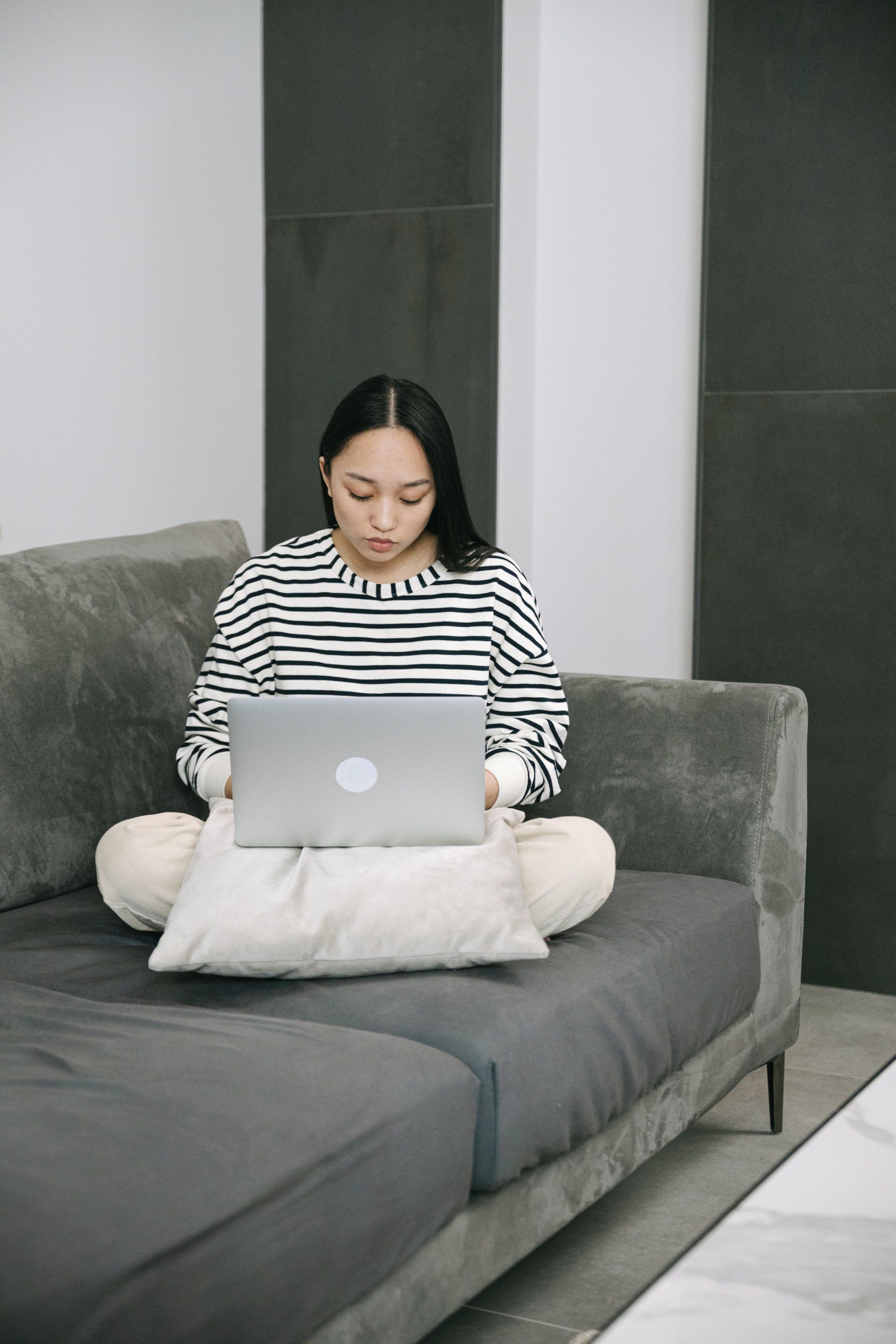 Woman on a gray sofa, working on a laptop. She is wearing a striped shirt and light pants.