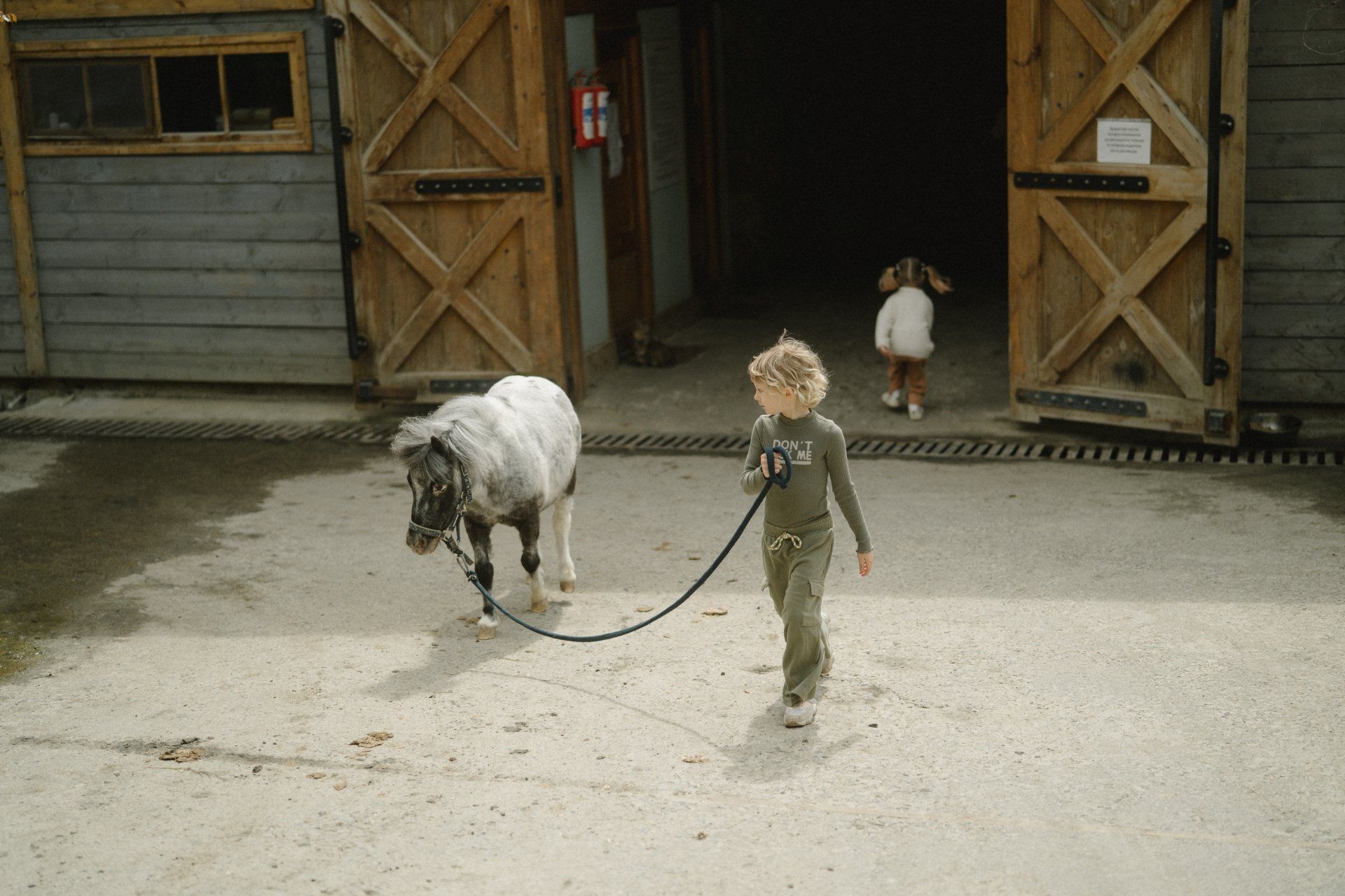 Een kind leidt een pony uit een schuur, een ander kind rent vooruit.