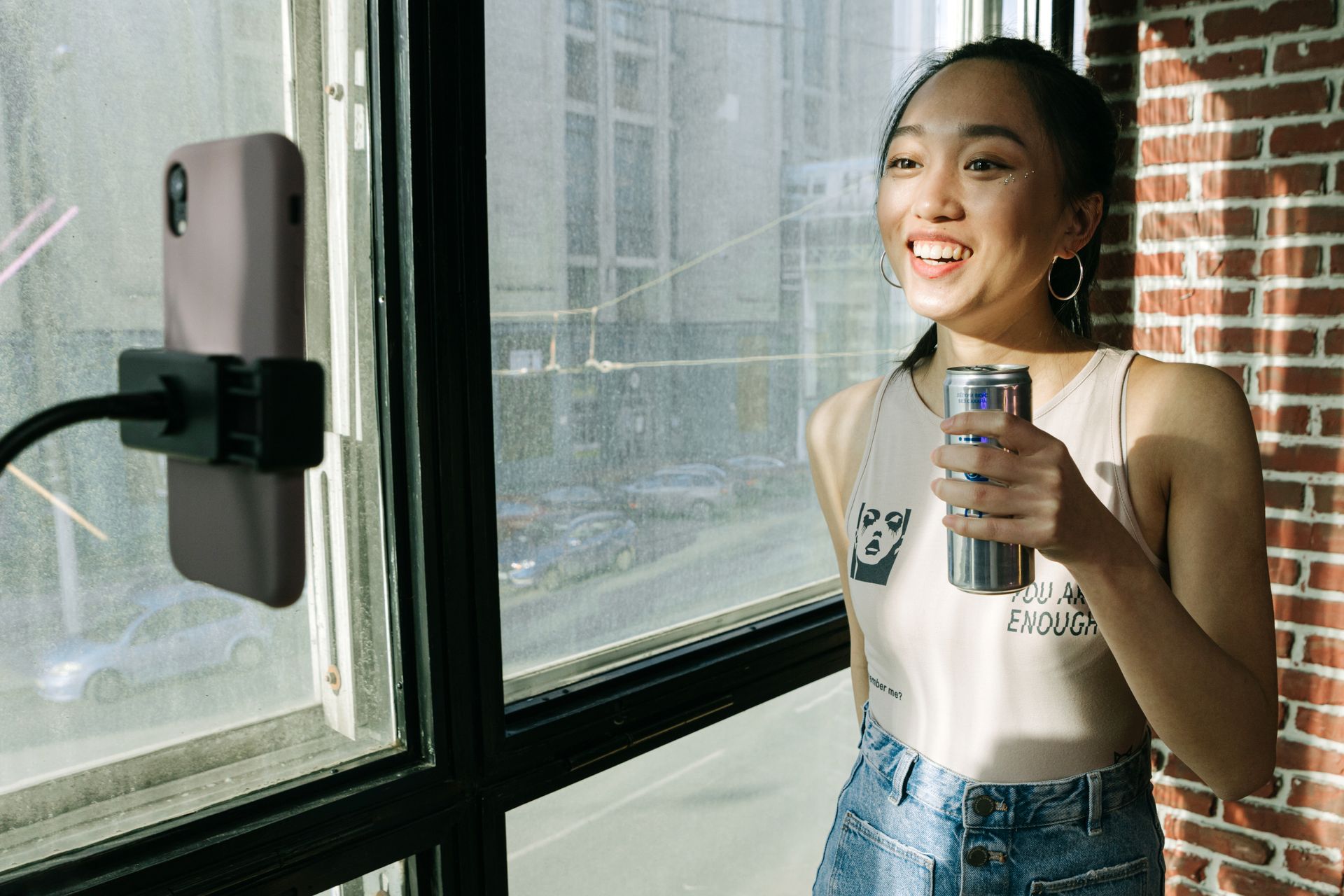 A woman is standing in front of a window holding a can of red bull and a cell phone.
