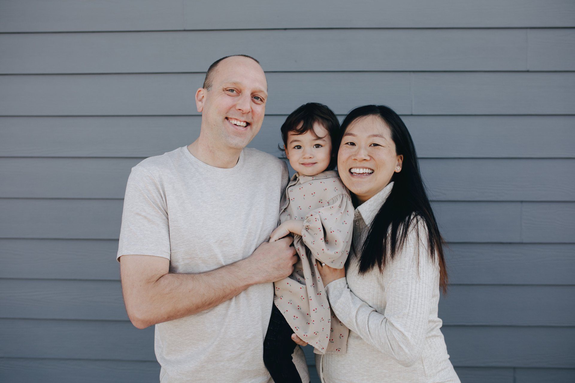 Three people smiling and holding a small dog in front of a gray wall.
