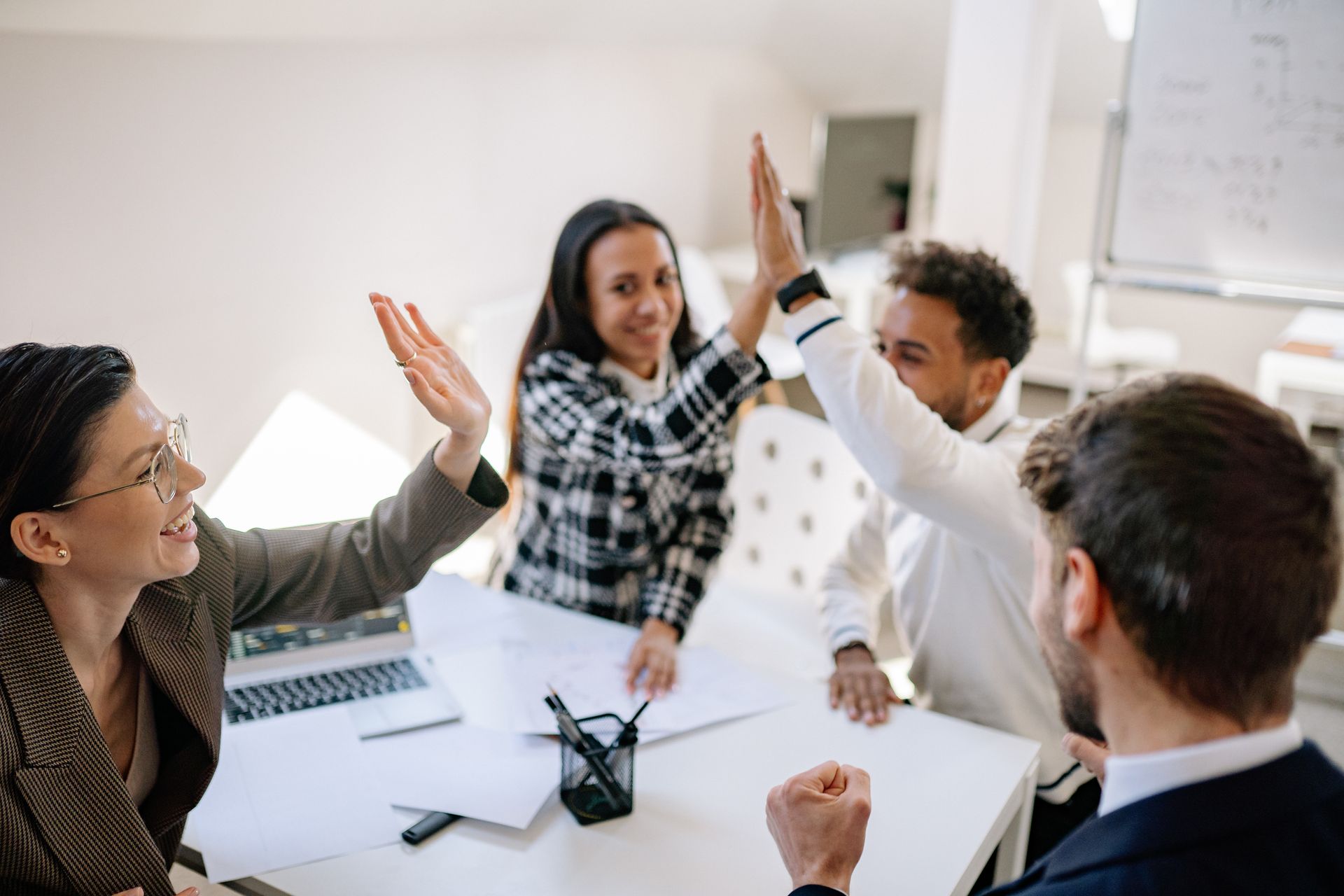 Four diverse colleagues high-fiving at a table in a bright office, celebrating success.