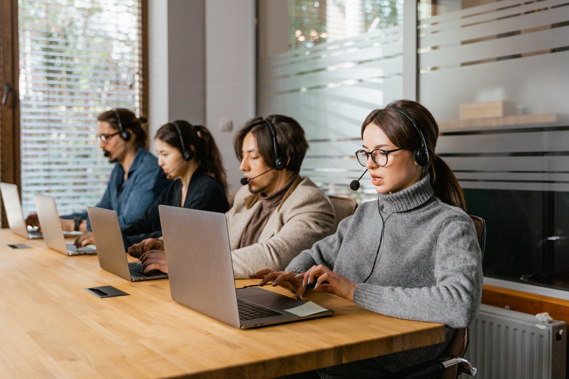 Four people with headsets working on laptops at a wooden table in an office setting.