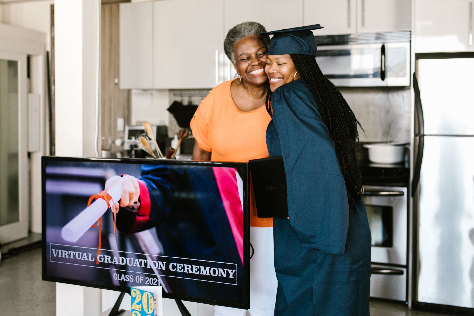 Woman in graduation gown hugs smiling woman in front of a TV displaying a virtual graduation ceremony.