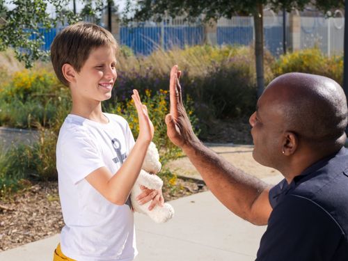 Boy and man high-five outdoors on a sunny sidewalk, smiling at each other.