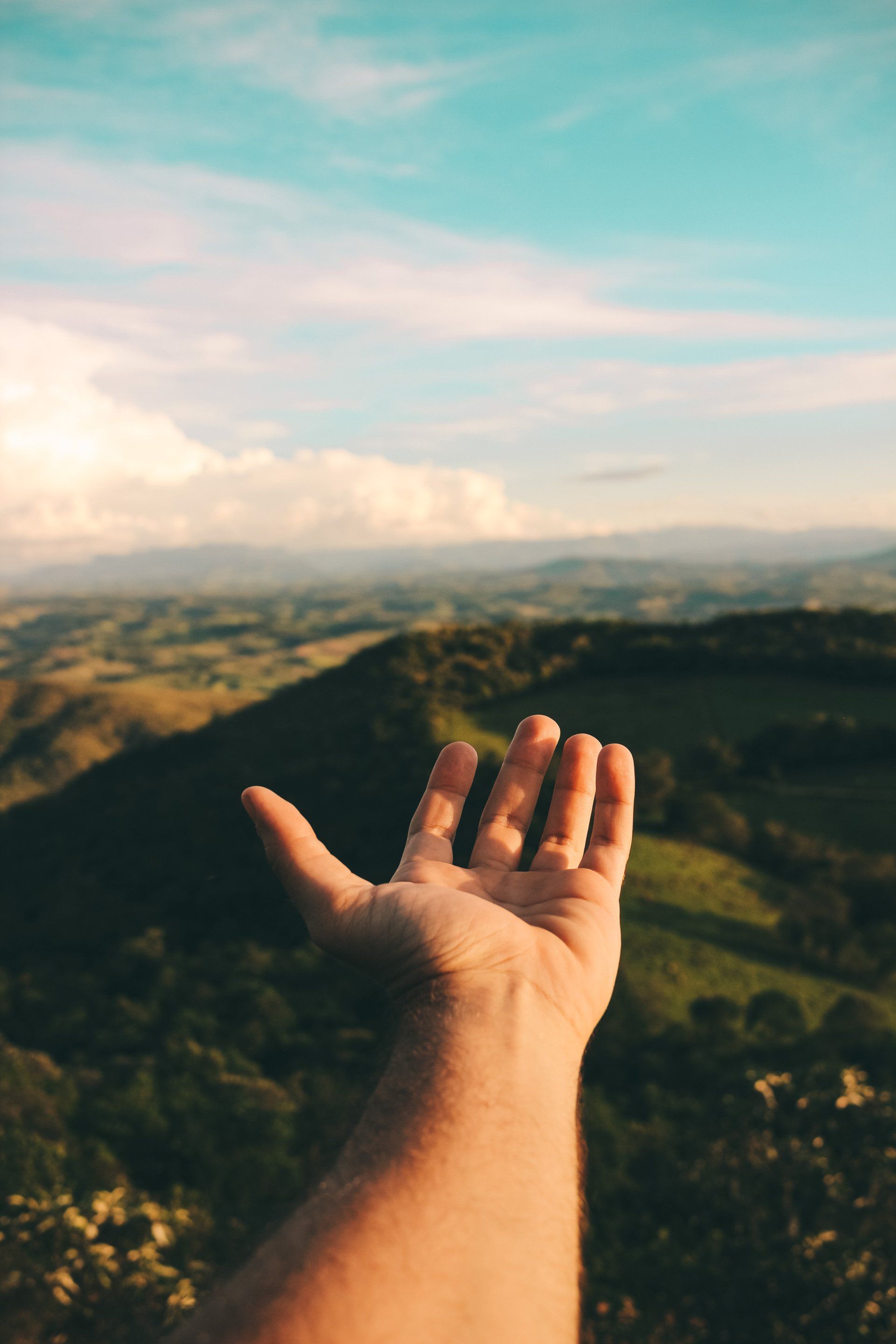 Outstretched hand reaching toward a sunlit mountain valley under a pastel sky