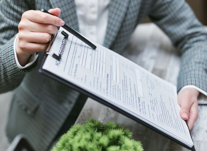 Woman in blazer points to a document on a clipboard, indoors.