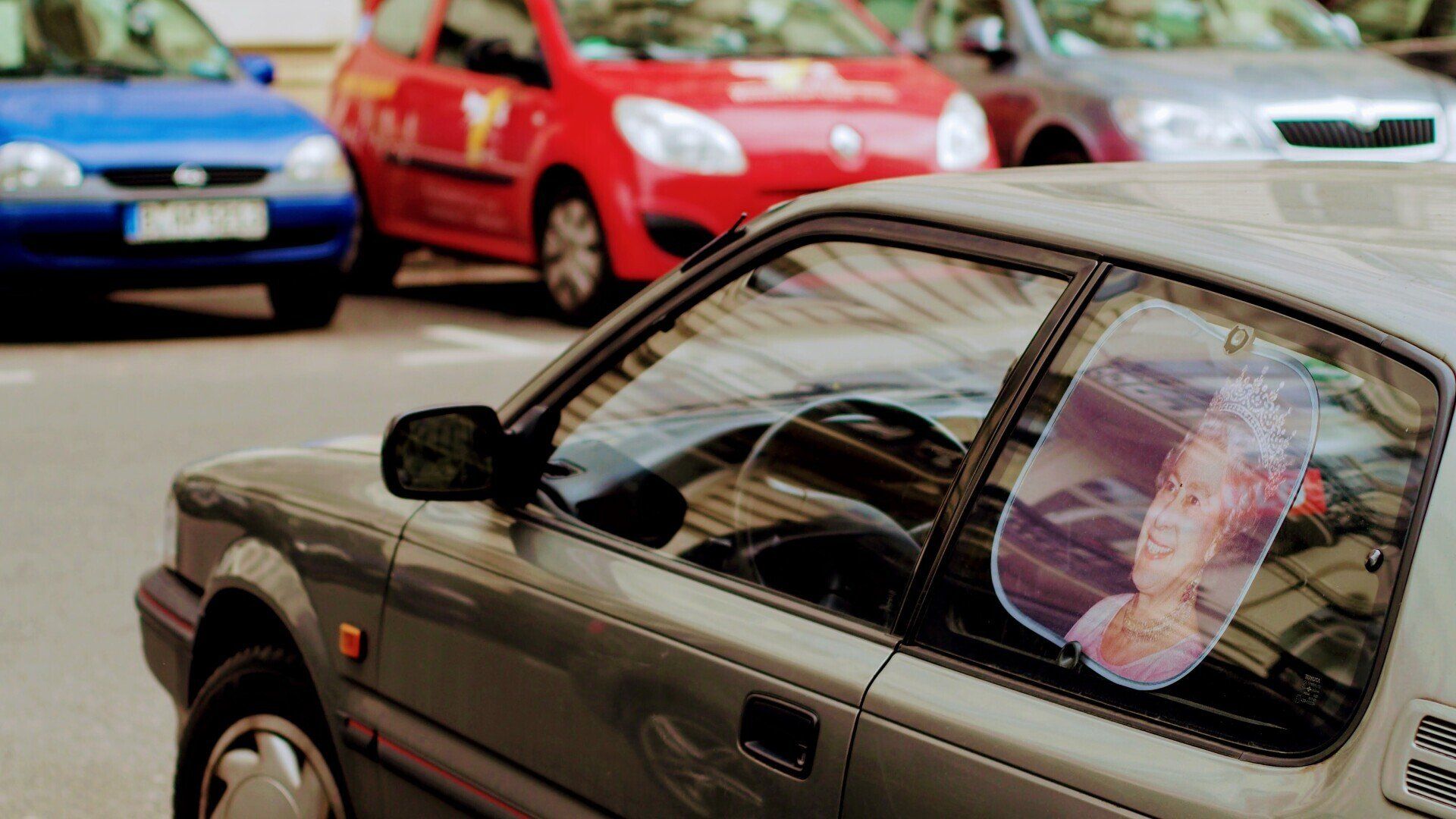 A gray car parked on a street with other cars; a picture is hanging in the car's window.