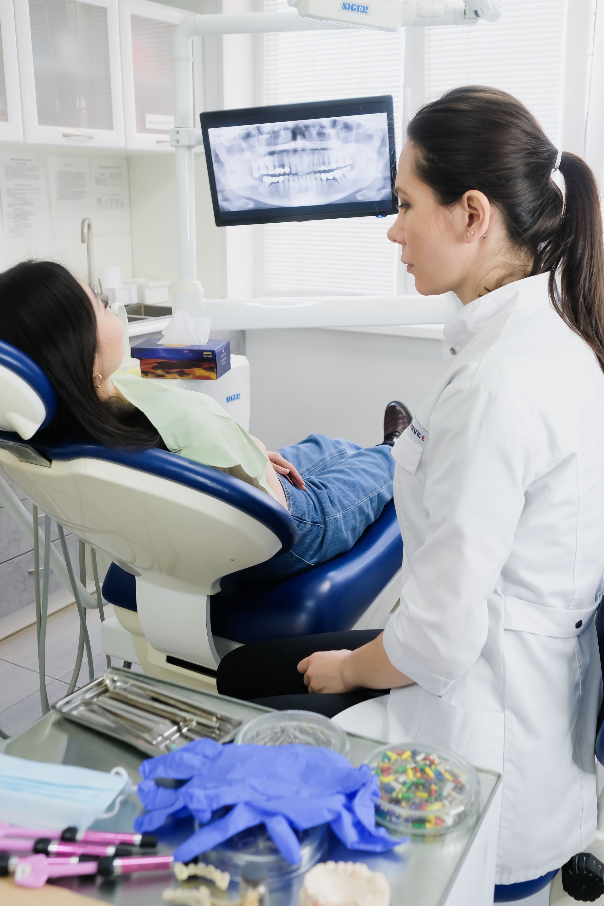 Dentist examining patient in a dental chair. X-ray on screen. Dental tools and supplies on table.