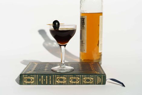 Cocktail glass with dark drink, olive garnish, on closed book; bottle of amber liquor behind it.