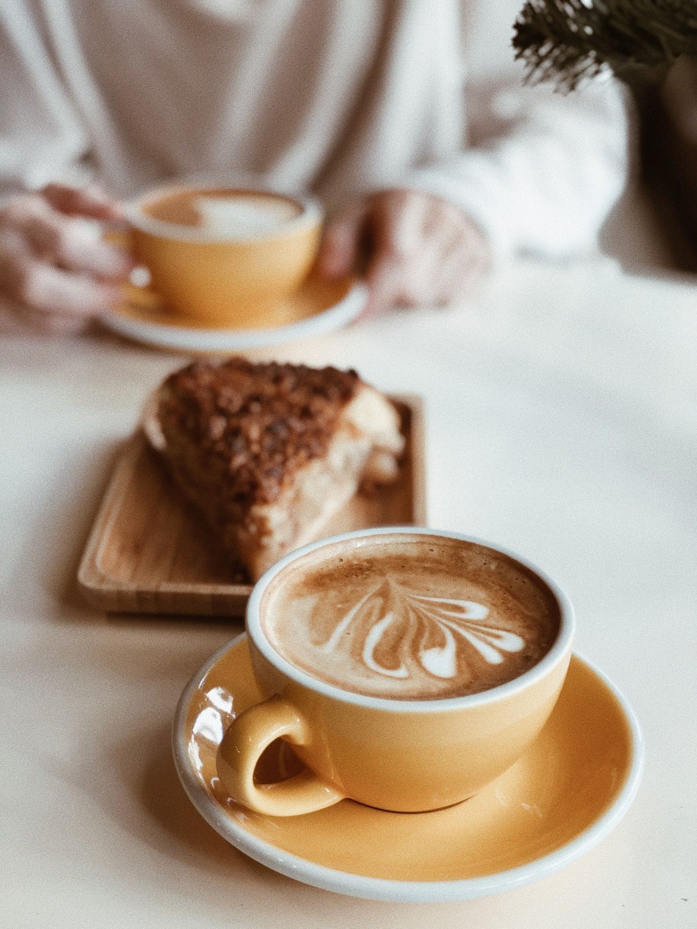 Two cups of latte on a table with a slice of cake on a wooden board, soft café setting