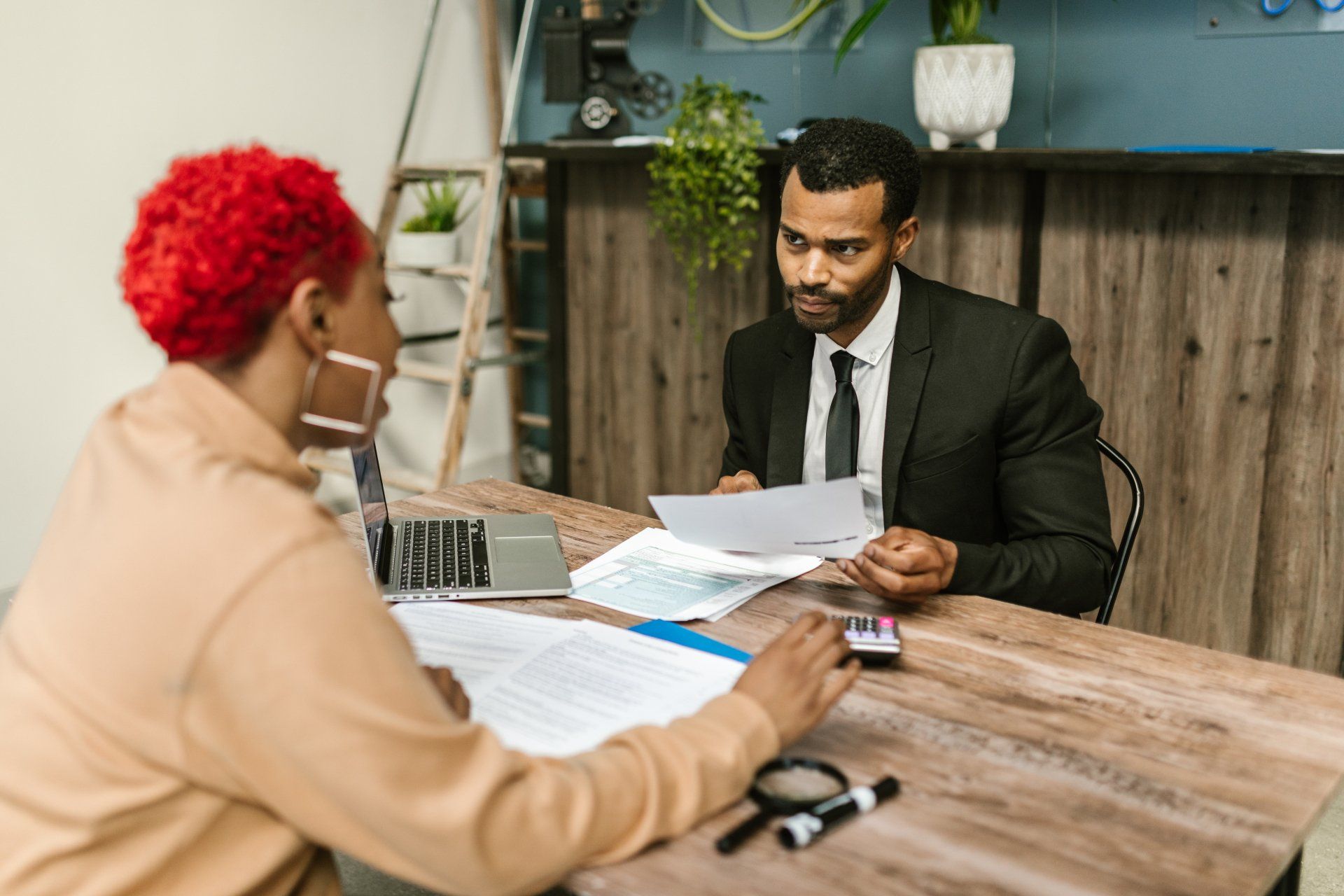 A man in a suit and tie is sitting at a table talking to a woman with red hair.