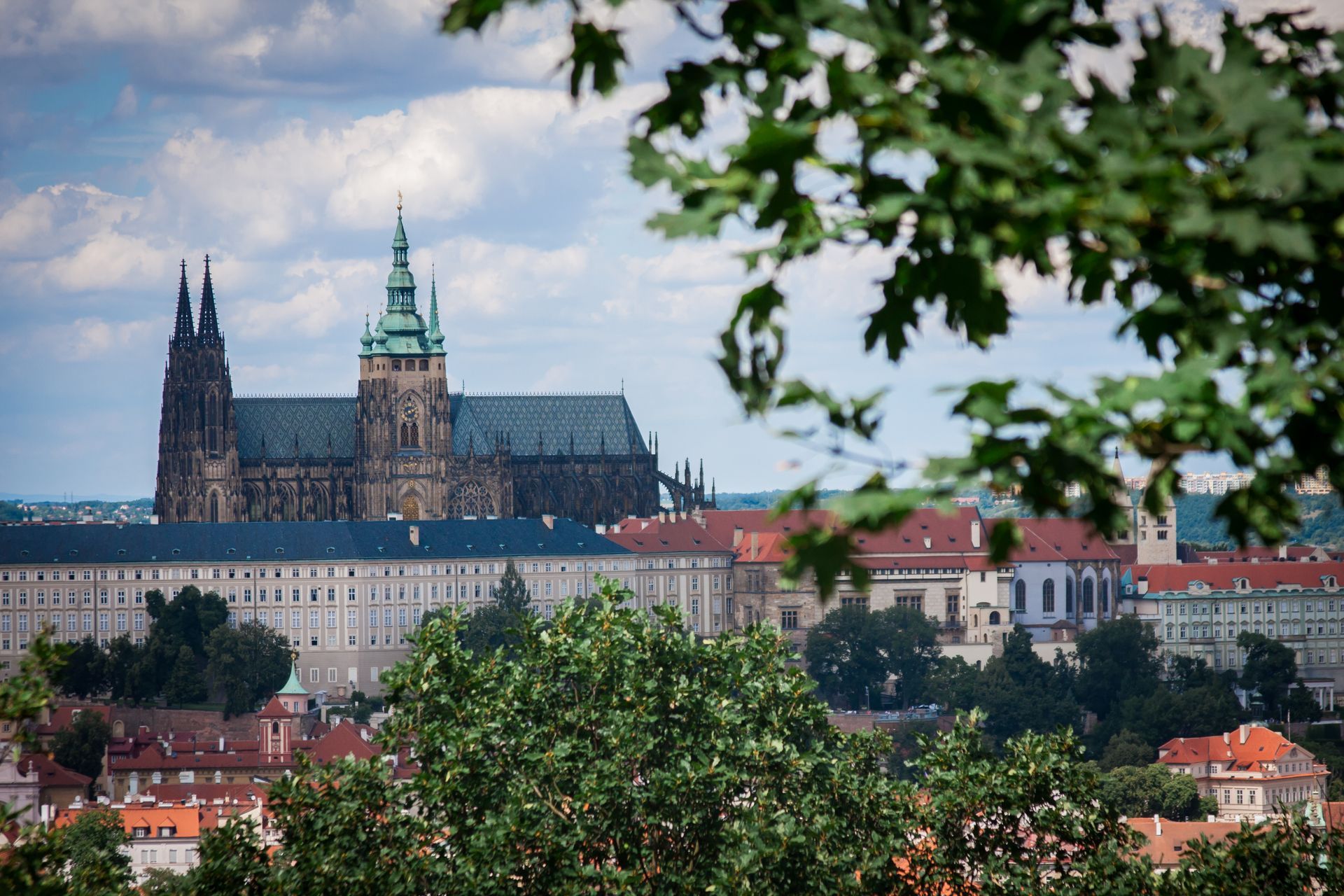 Historic cathedral and castle skyline above red-roofed houses, framed by green trees under a cloudy sky