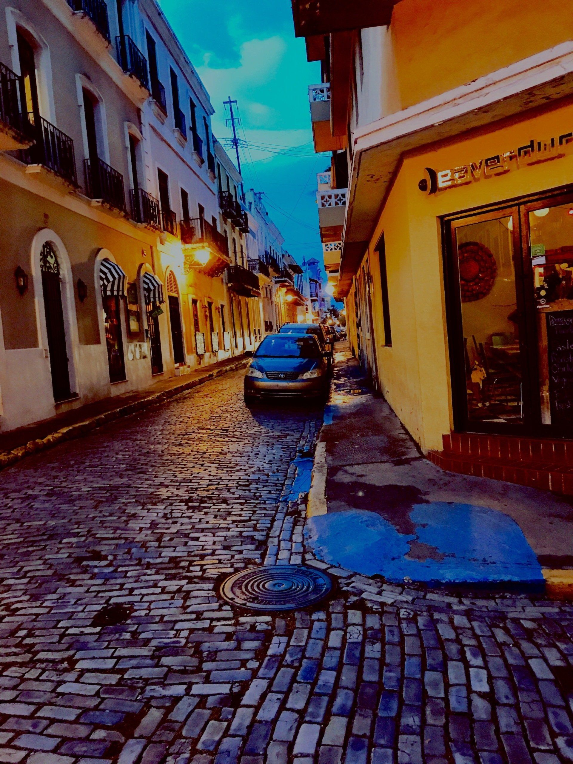 Cobblestone street in Old San Juan, Puerto Rico, lined with colorful buildings. A car is parked down the street.