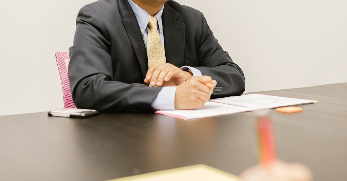 Businessperson seated at a desk with papers, hands clasped in a meeting room