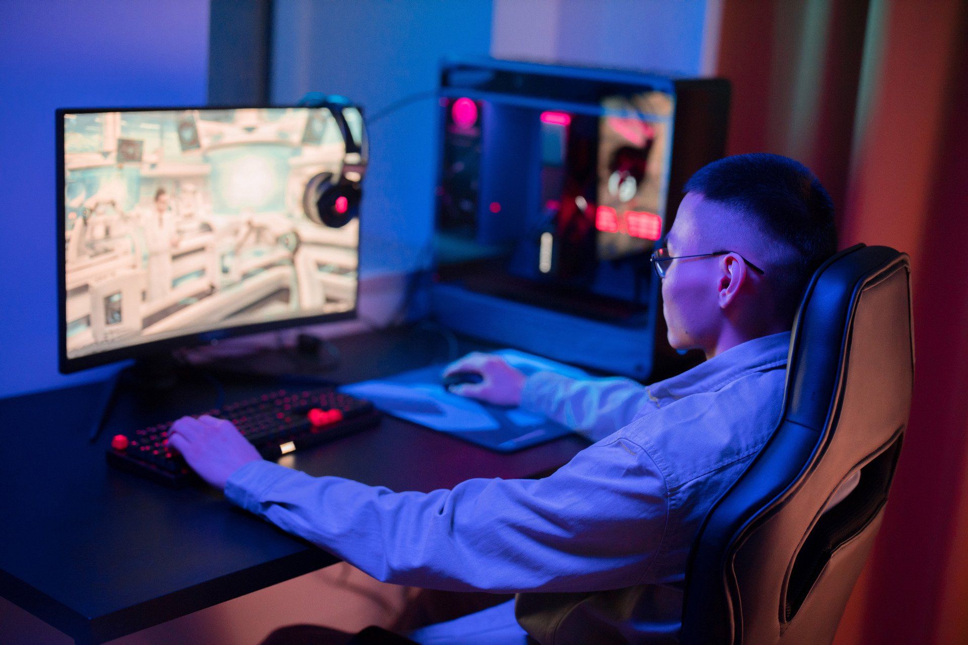 Person gaming on a computer, illuminated by blue light. Keyboard, monitor, PC tower, and headphones visible.