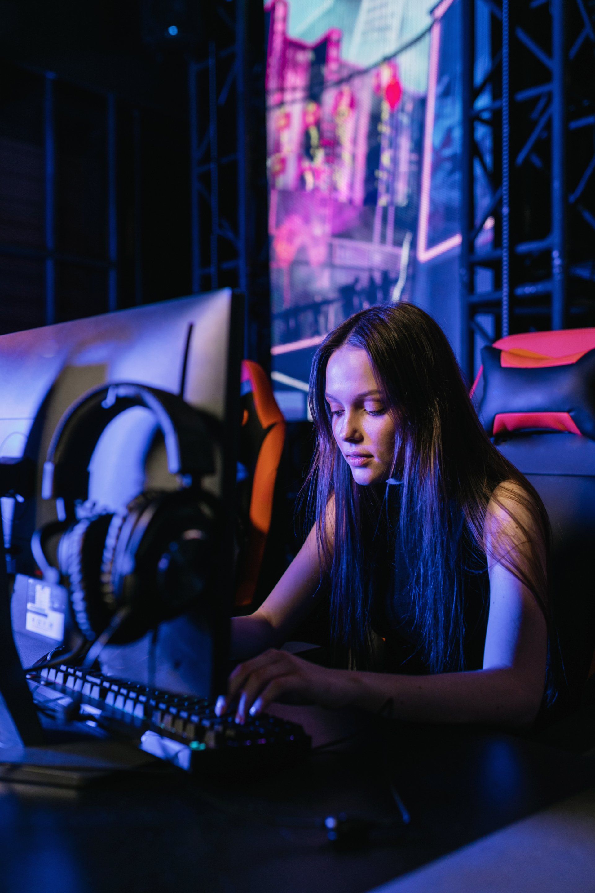 Woman typing on a keyboard in a gaming setup. Dark room with a large screen in the background.