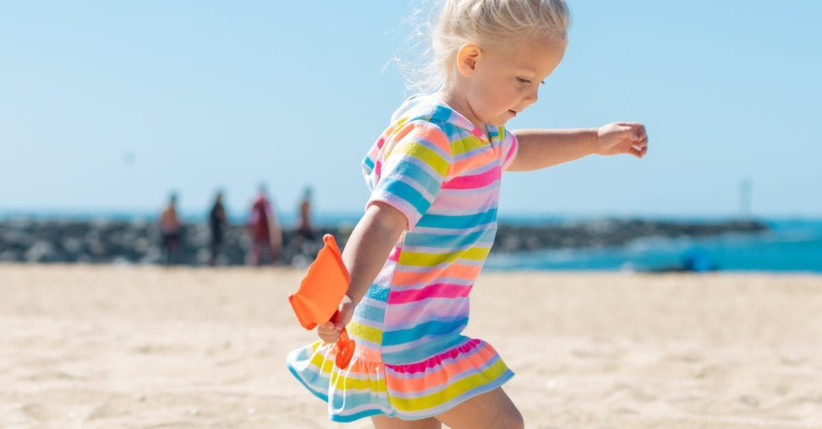 Criança de vestido listrado caminhando em uma praia de areia com o oceano ao fundo.