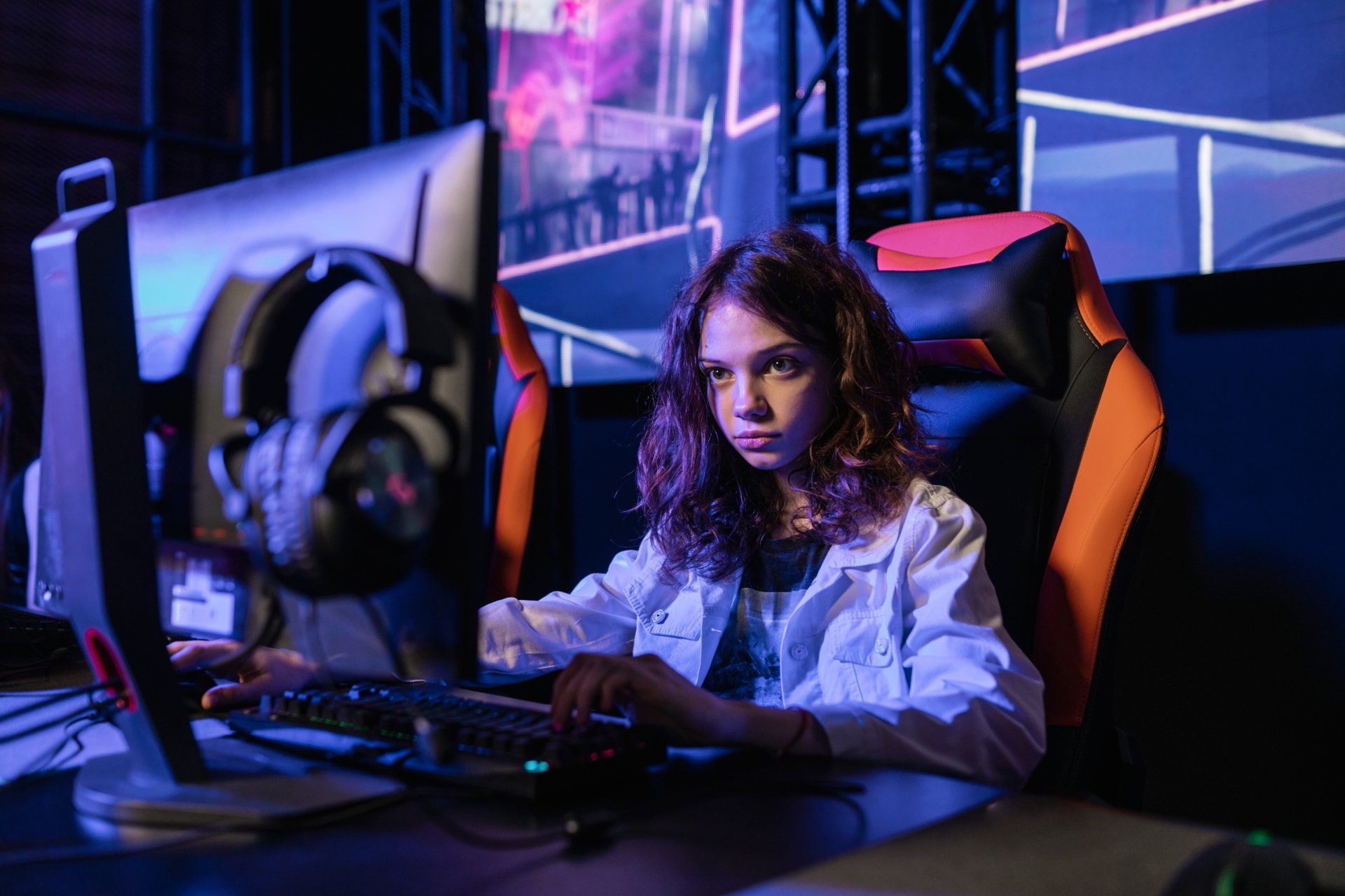 Young person intently using a computer in a gaming setup, with headphones and a backlit screen.