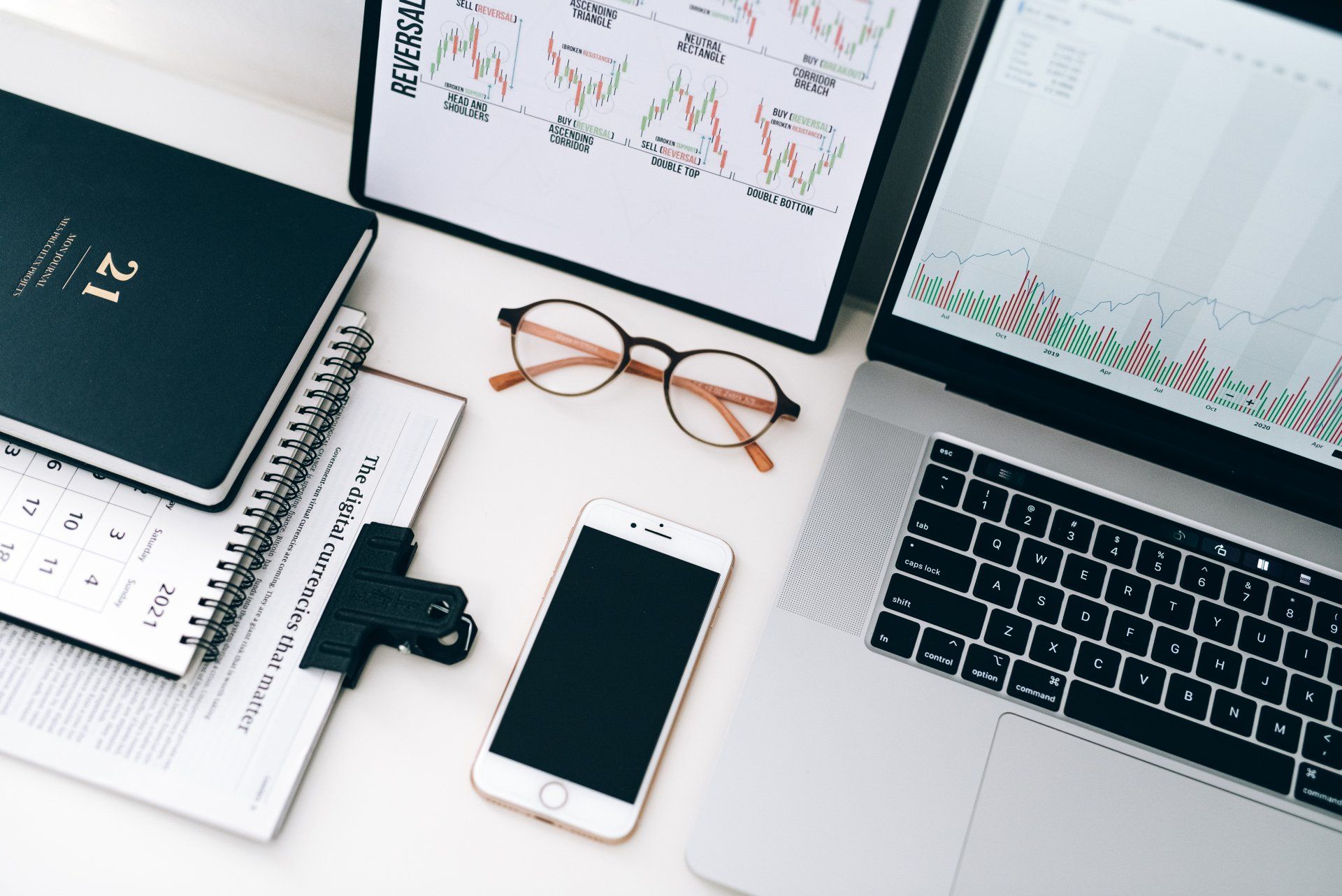 Laptop, phone, eyeglasses, notebook, and financial charts on a white desk.