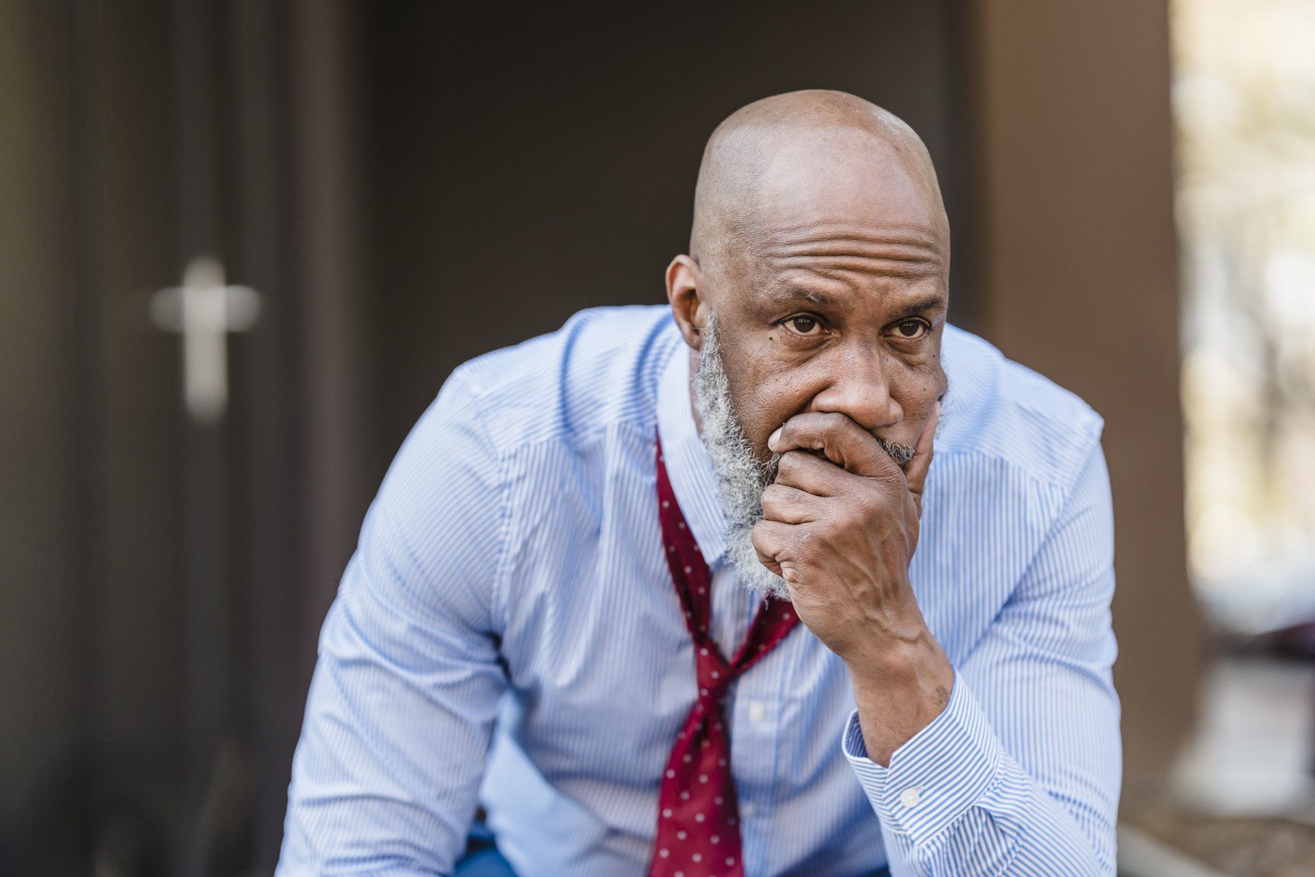 A man in a blue shirt and red tie is sitting down with his hand on his chin.