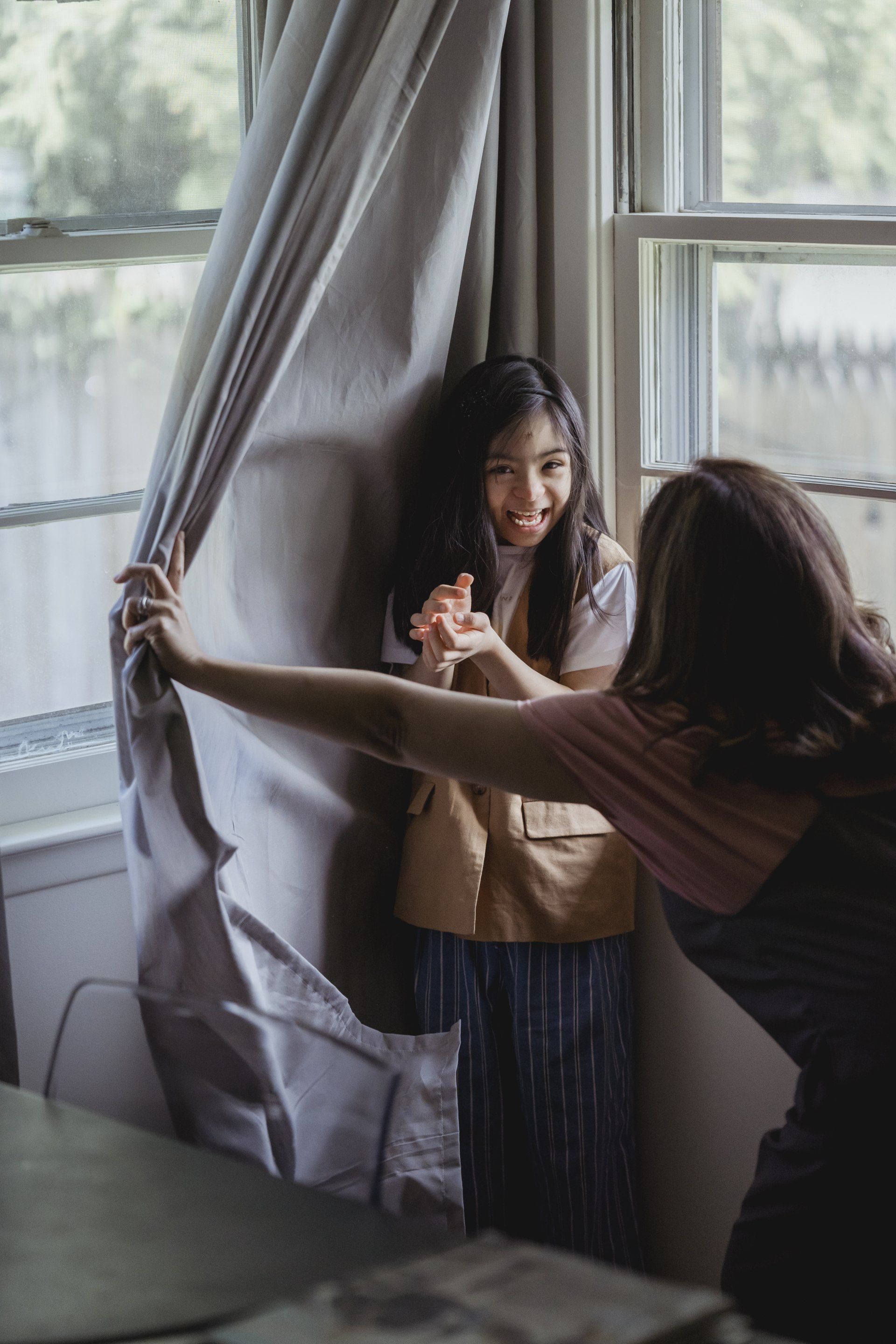 A woman is holding a child 's hand while opening a window curtain.