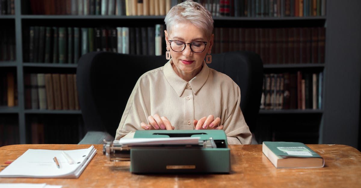 An elderly woman is typing on a typewriter at a desk in a library.