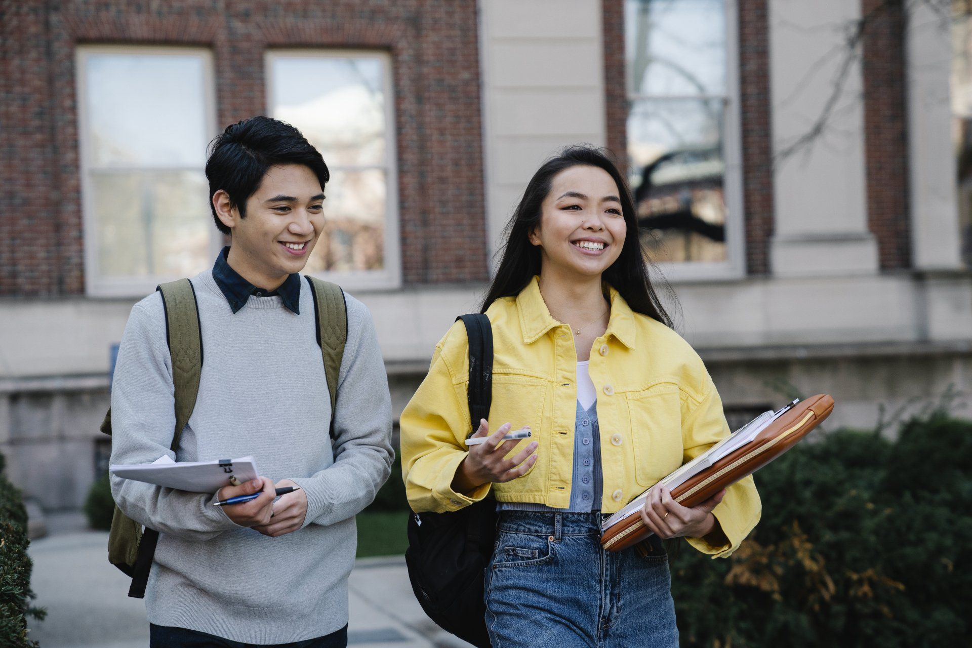 Two students walking and smiling in front of a brick building, one holding a tablet and the other holding a notebook.